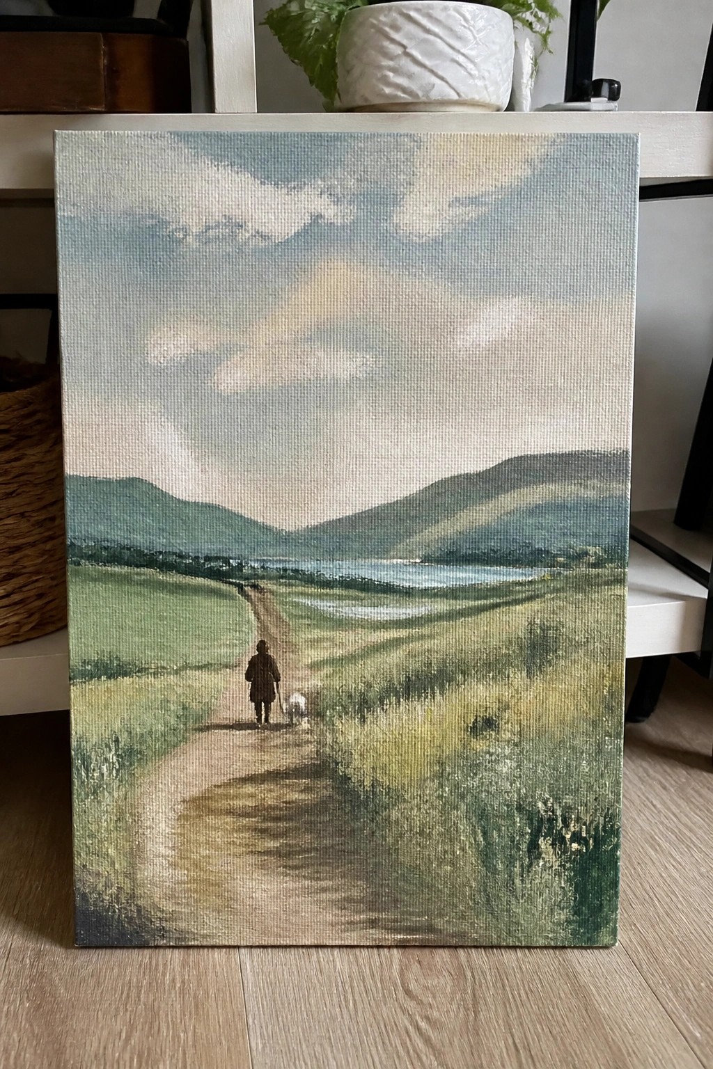 Landscape painting of person walking dog on path through green fields toward hills and lake.