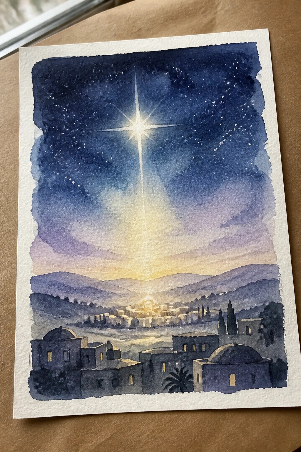 Watercolor night sky with radiant star glowing over domed town and hills.