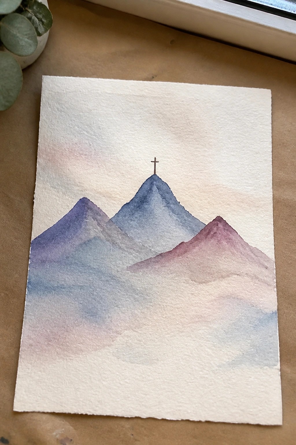 Watercolor painting of misty blue-purple mountains with cross atop central peak