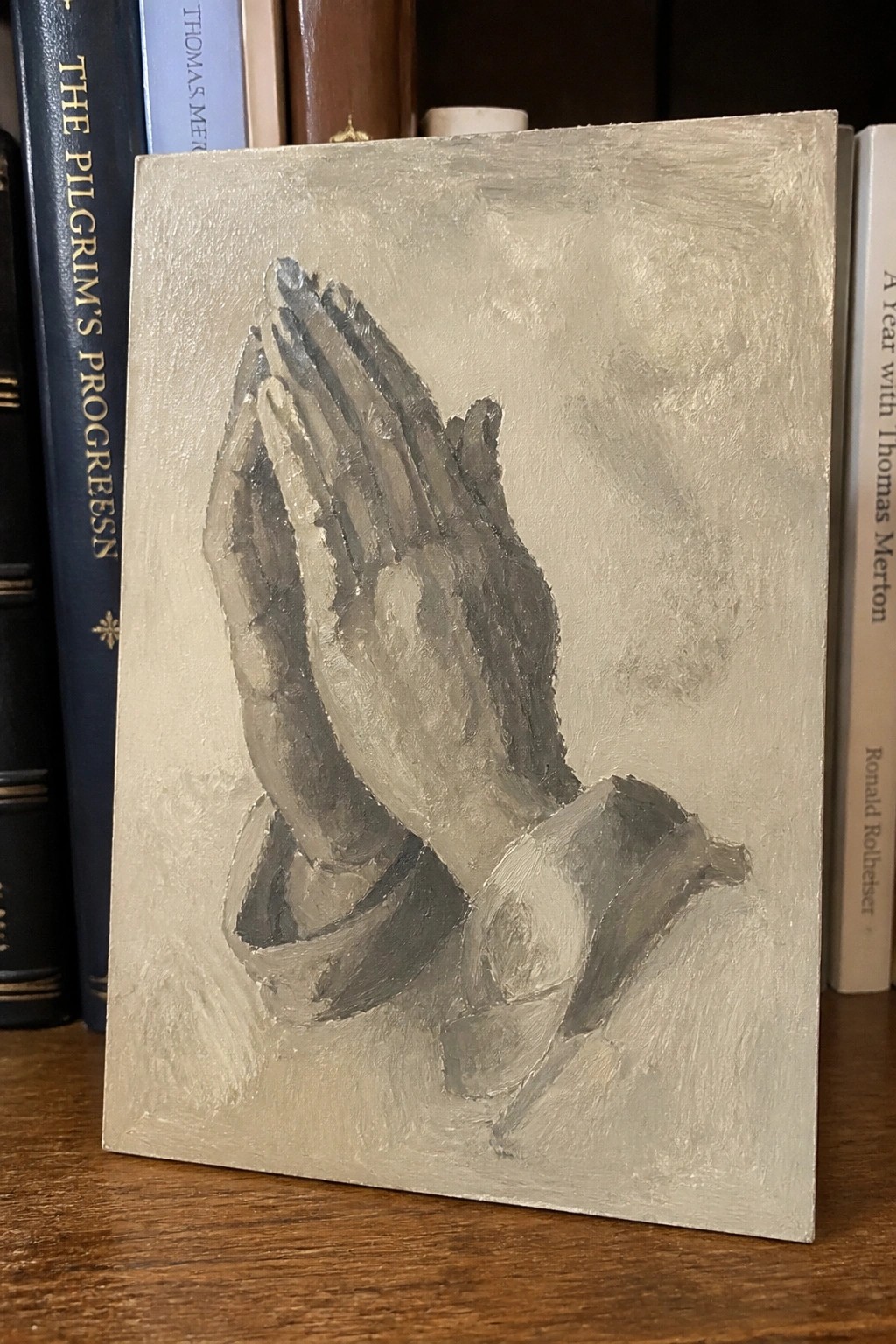 Painted praying hands in grayscale on small canvas beside books