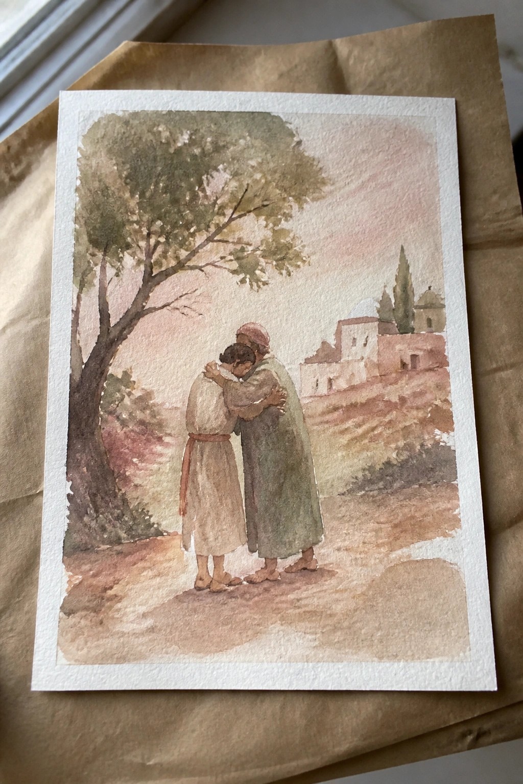 Two robed figures embrace on a dirt path by a tree with distant buildings in watercolor.