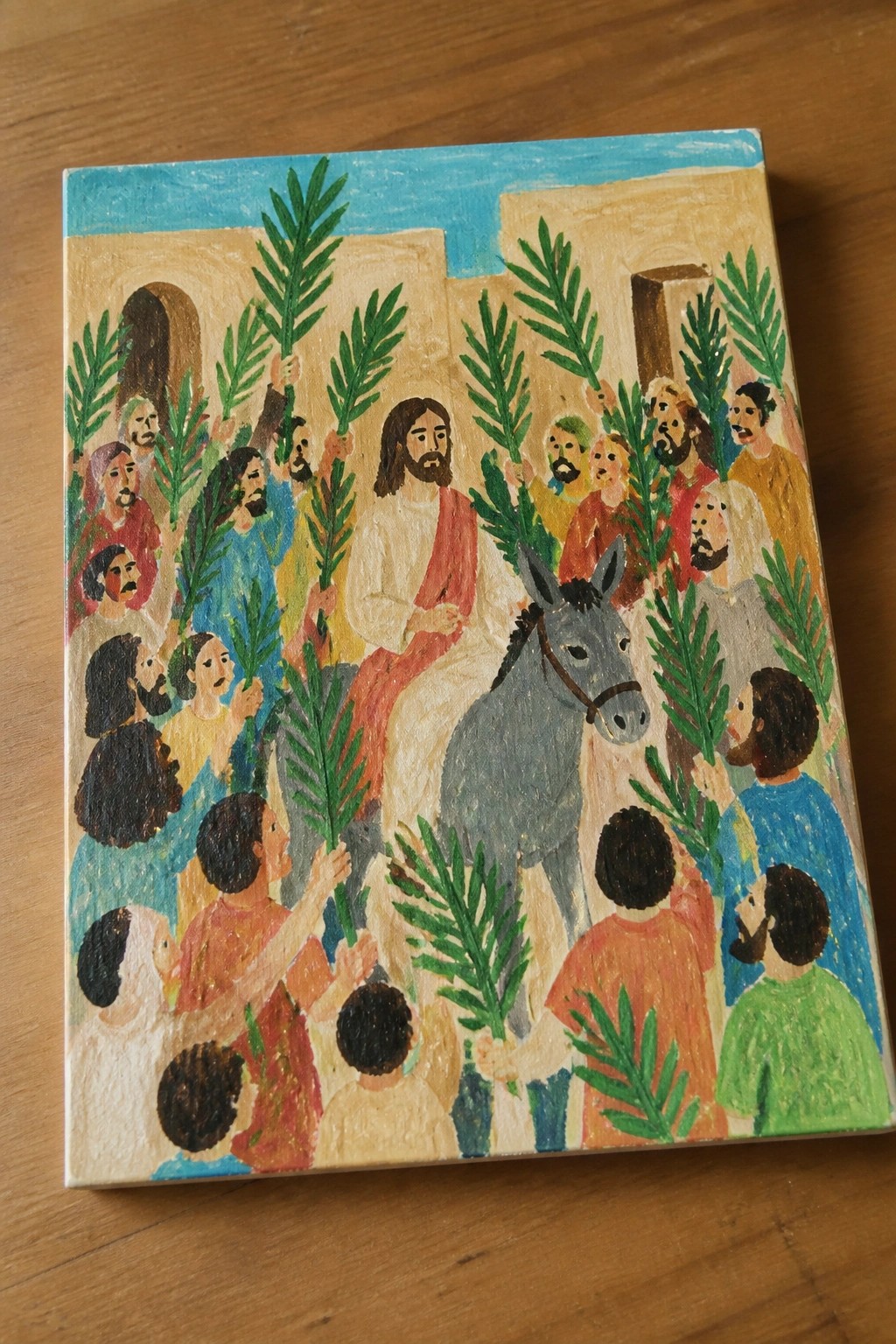 Colorful folk-art painting of Jesus on gray donkey amid crowd waving green palm fronds.