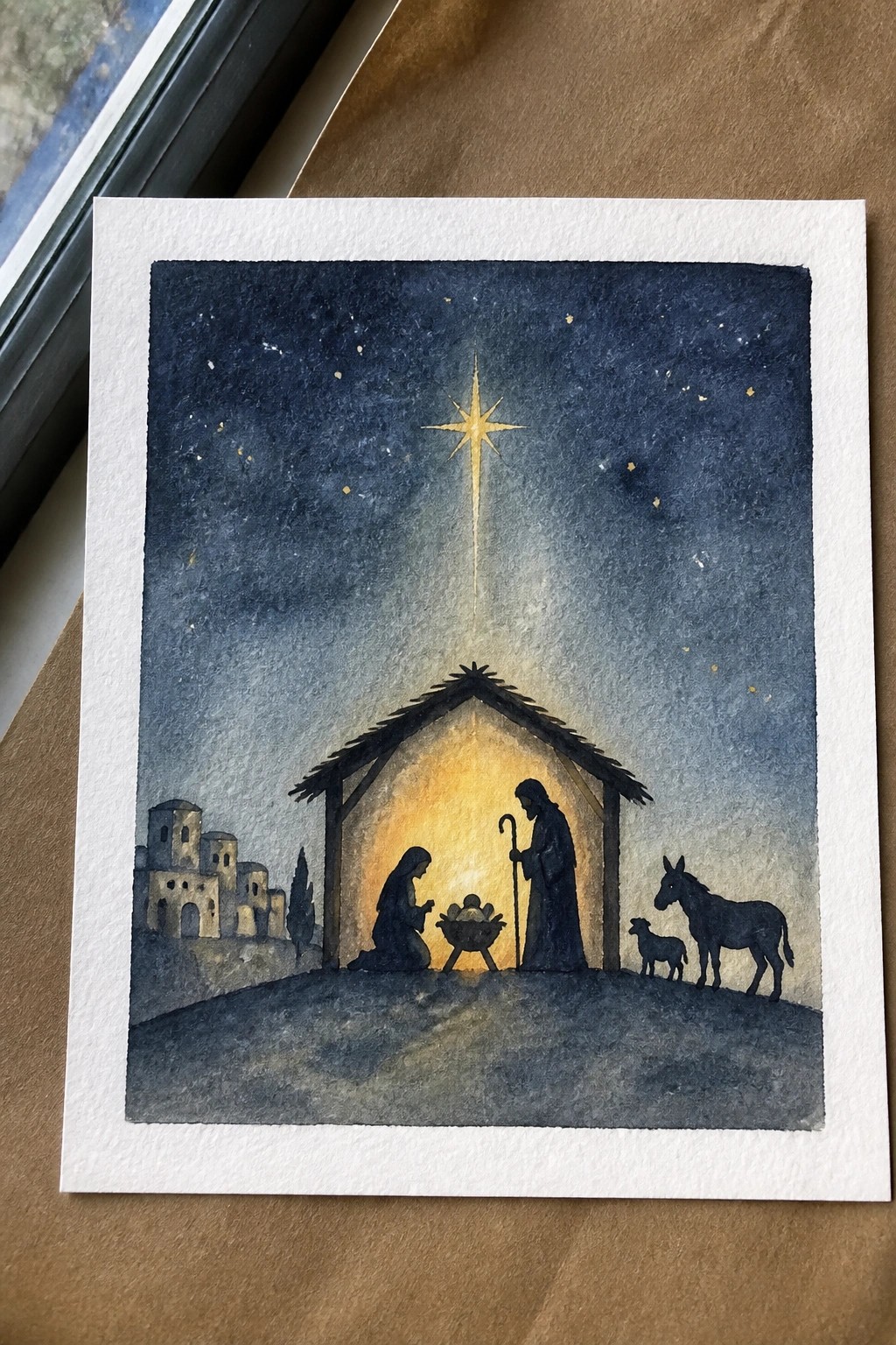Watercolor nativity with silhouetted Mary, Joseph, manger, donkey and lamb under glowing star.