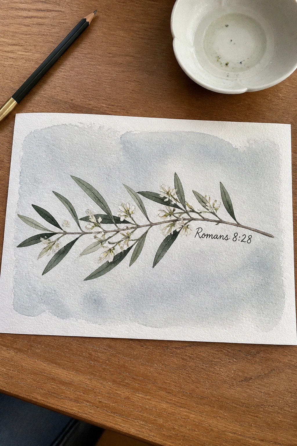 Watercolor olive branch with flowers and Romans 8:28 on paper beside pencil and bowl on wood