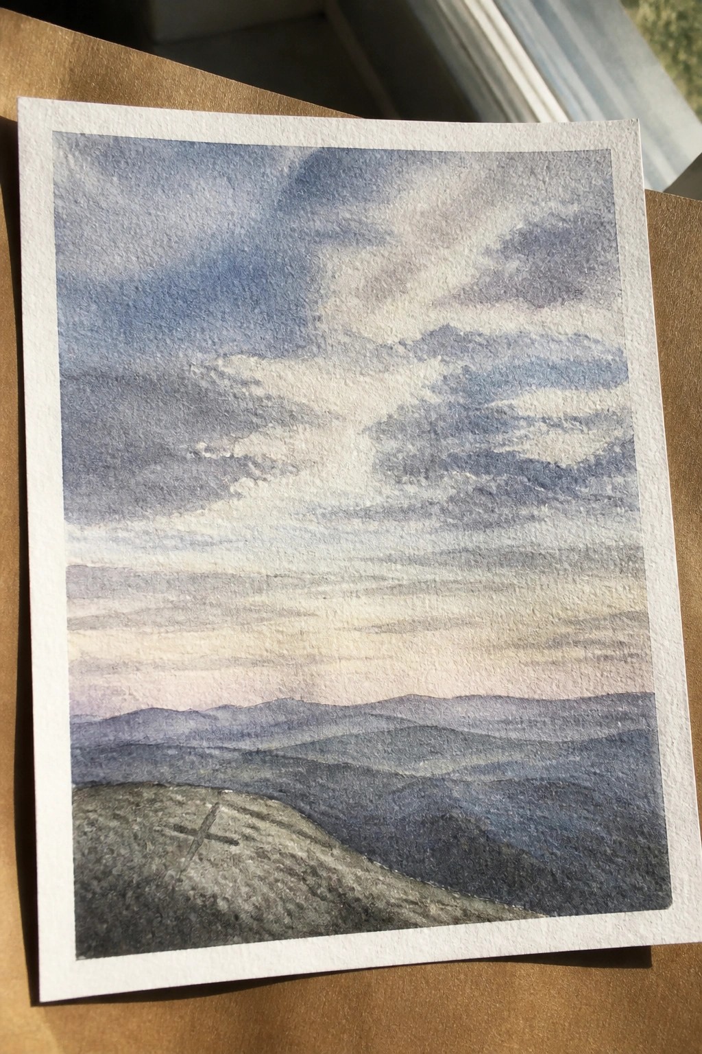 Watercolor of layered blue hills under cloudy sky, small cross on foreground slope.