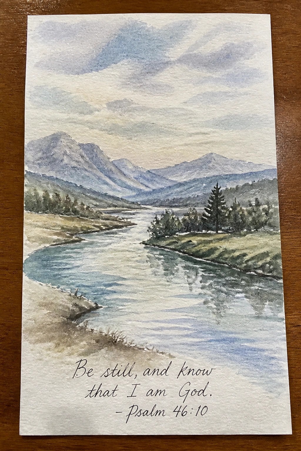 Watercolor landscape of a winding river through mountains with Psalm 46:10 text at the bottom.