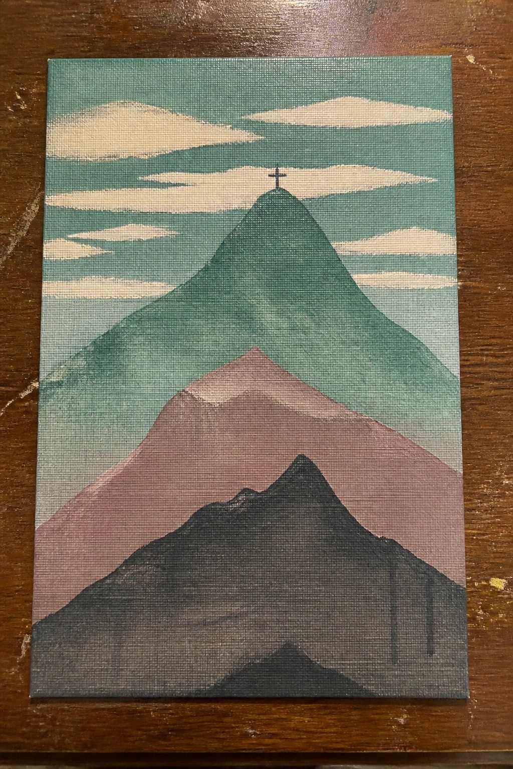 Layered mountains in green, pink, and gray with cross on tallest peak under cloudy sky.
