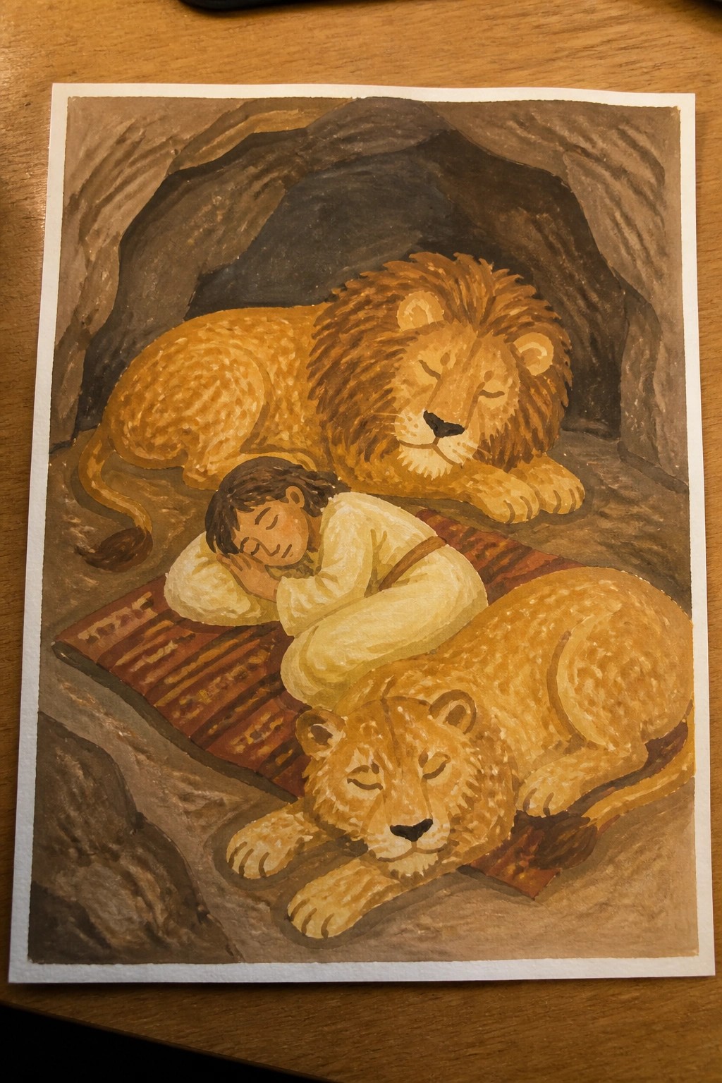 A watercolor painting shows a boy in light clothing sleeping on a patterned rug inside a cave, with a large lion behind him and a smaller lion in front, both lions resting with eyes closed.
