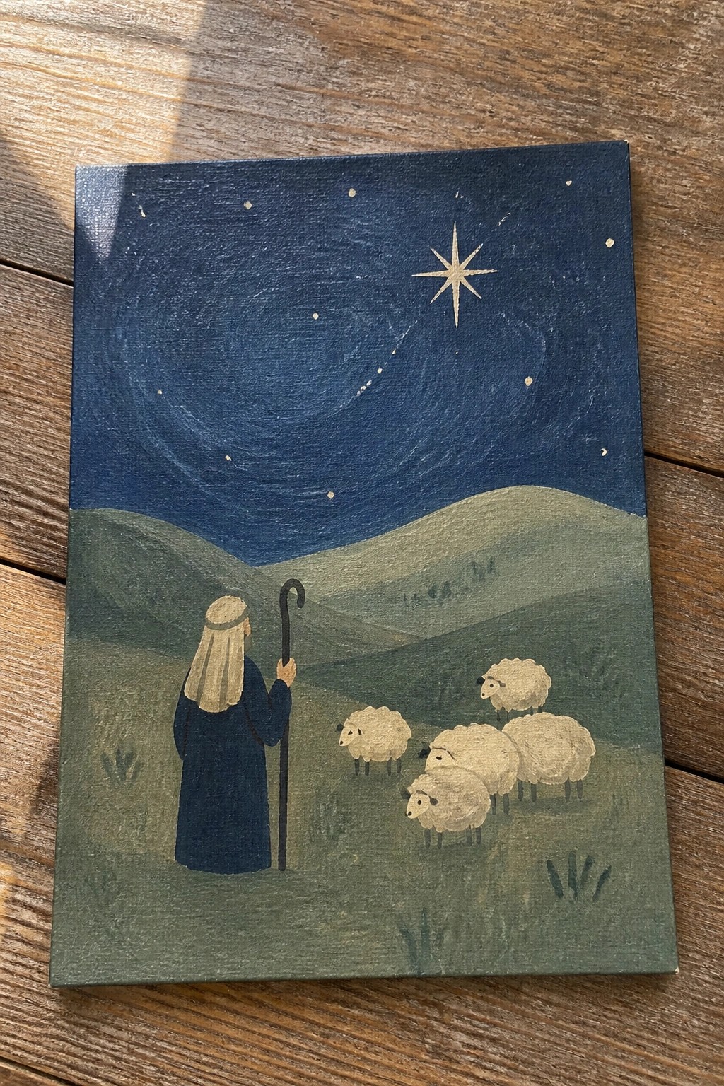 Shepherd in blue robe with staff watches sheep under large star in night sky