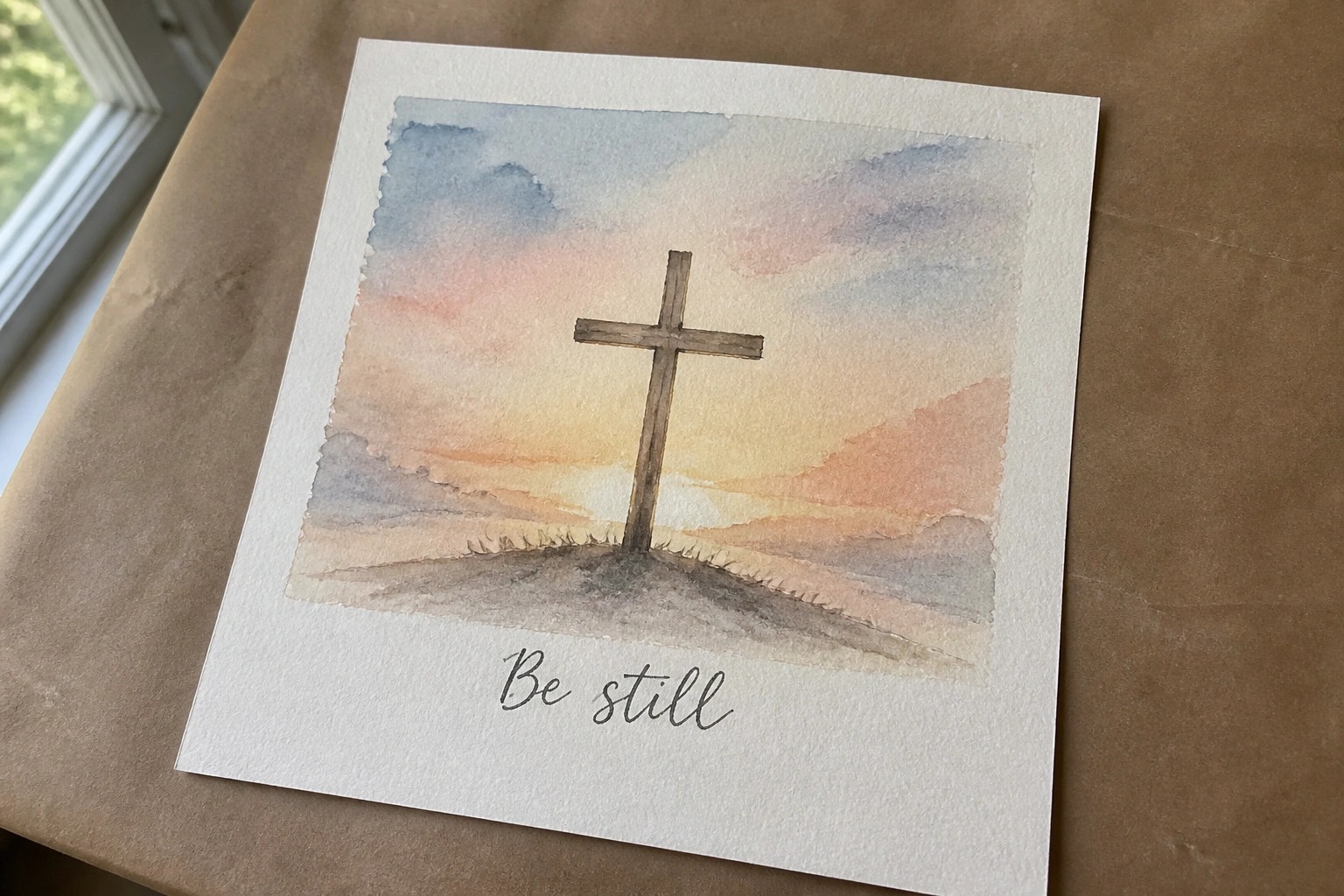Watercolor painting of wooden cross on hill at sunset with "Be still" text