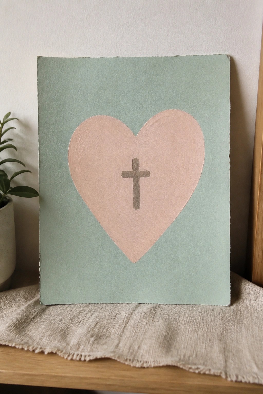 Pink heart with gray cross on mint green canvas atop beige cloth.