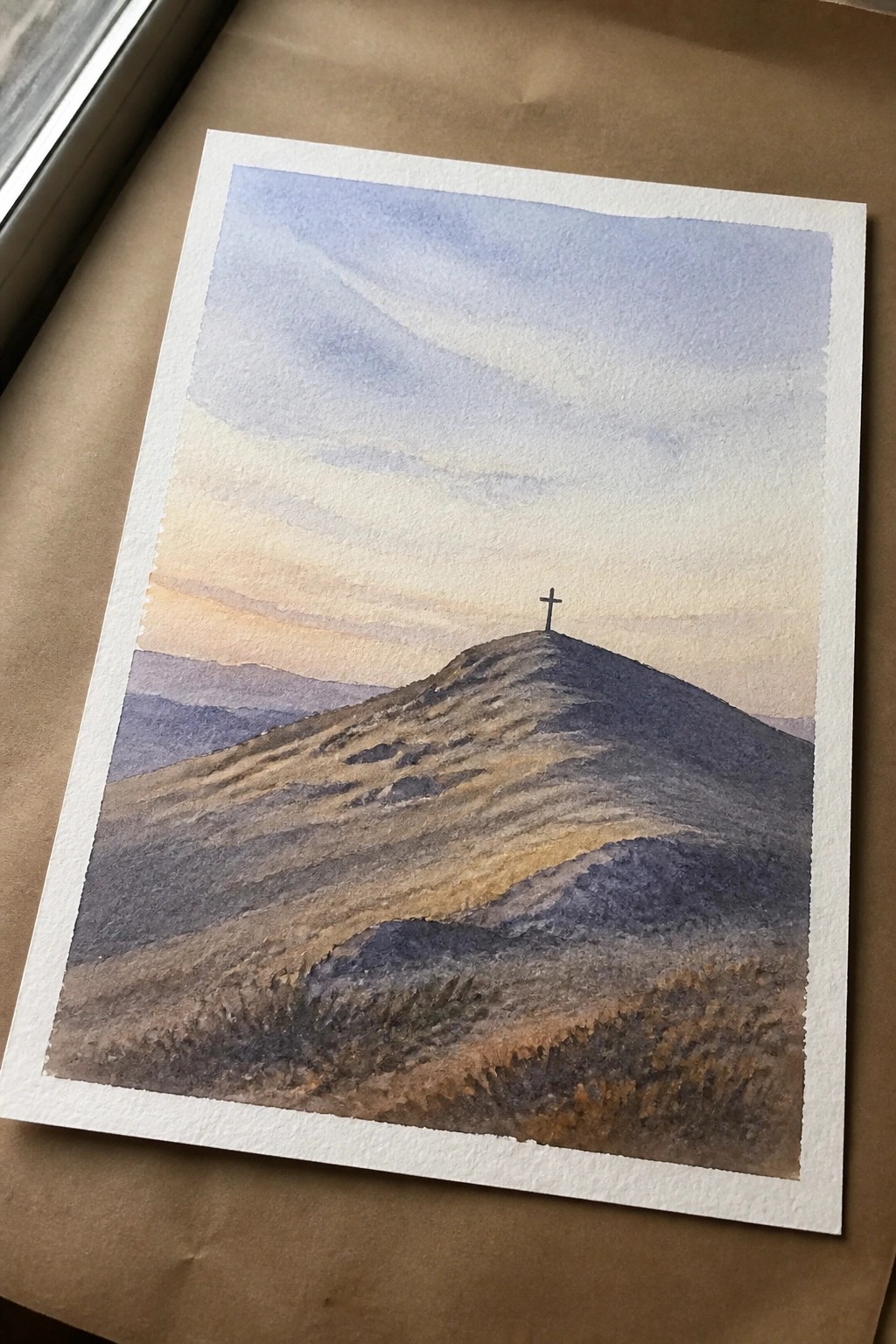 A watercolor painting of a hill with a cross on top under a sunset sky with layered mountains in the background.