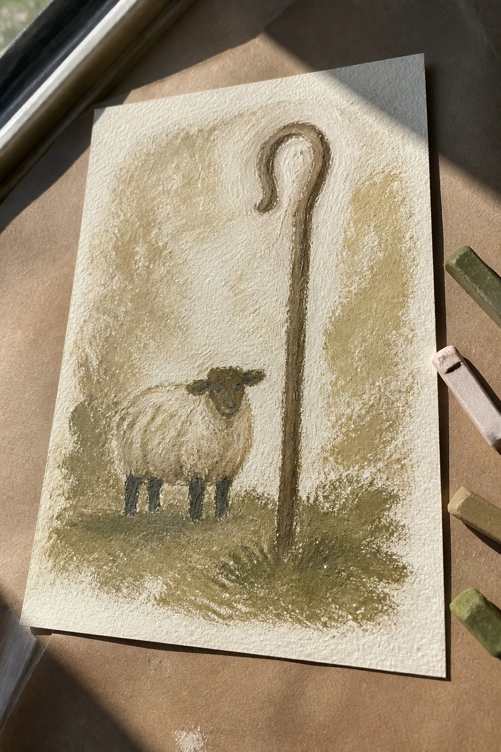 Pastel drawing of white sheep with black legs beside tall brown shepherd's staff on paper