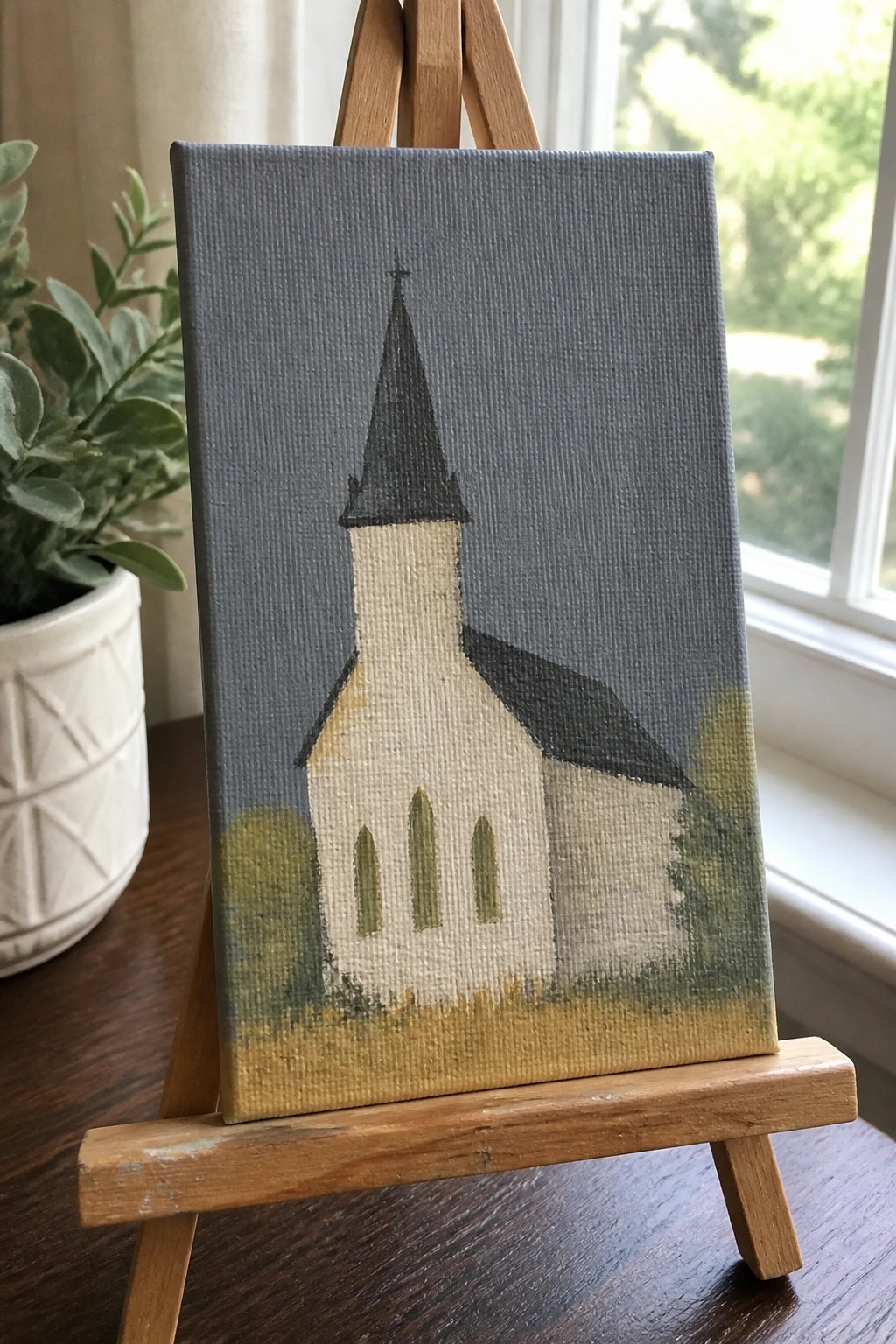 Small white church painting on wooden easel beside potted plant and window