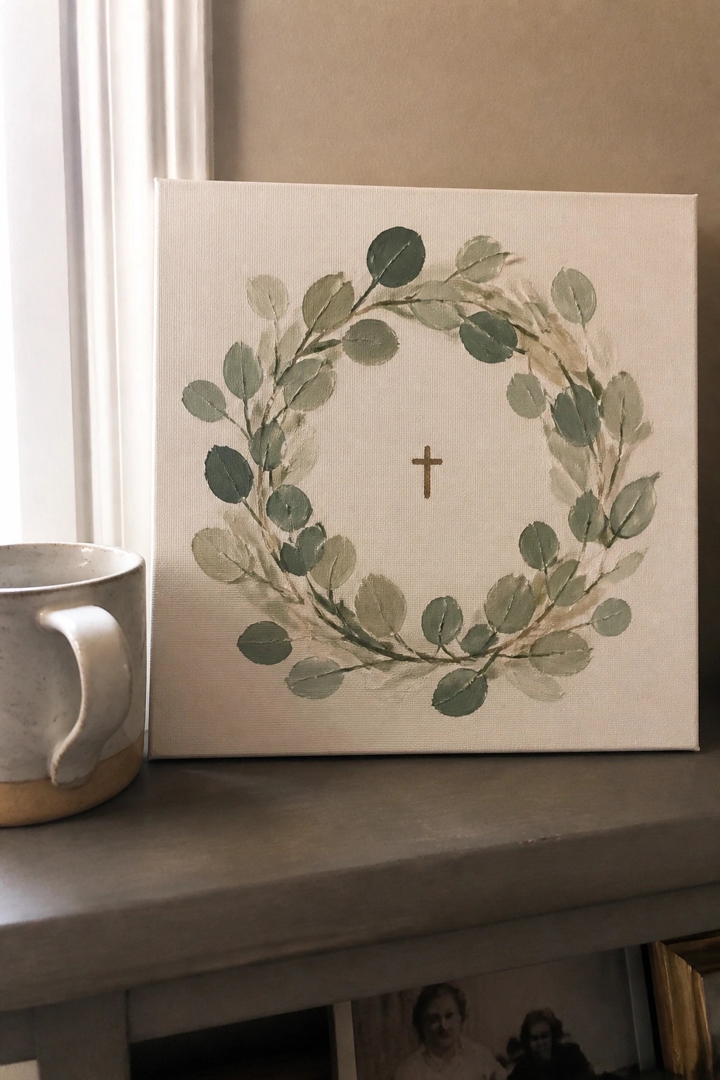 Sage green eucalyptus wreath with cross painted on canvas beside ceramic mug