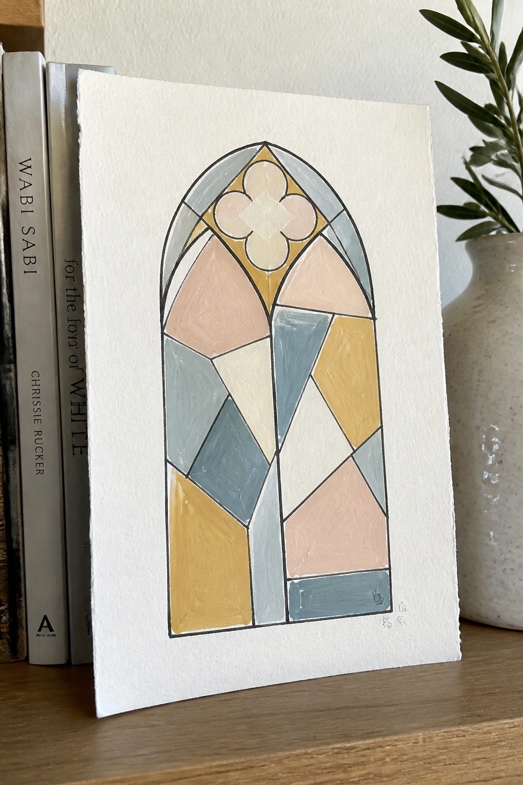Pastel arched stained glass painting on paper beside books and ceramic vase with plant