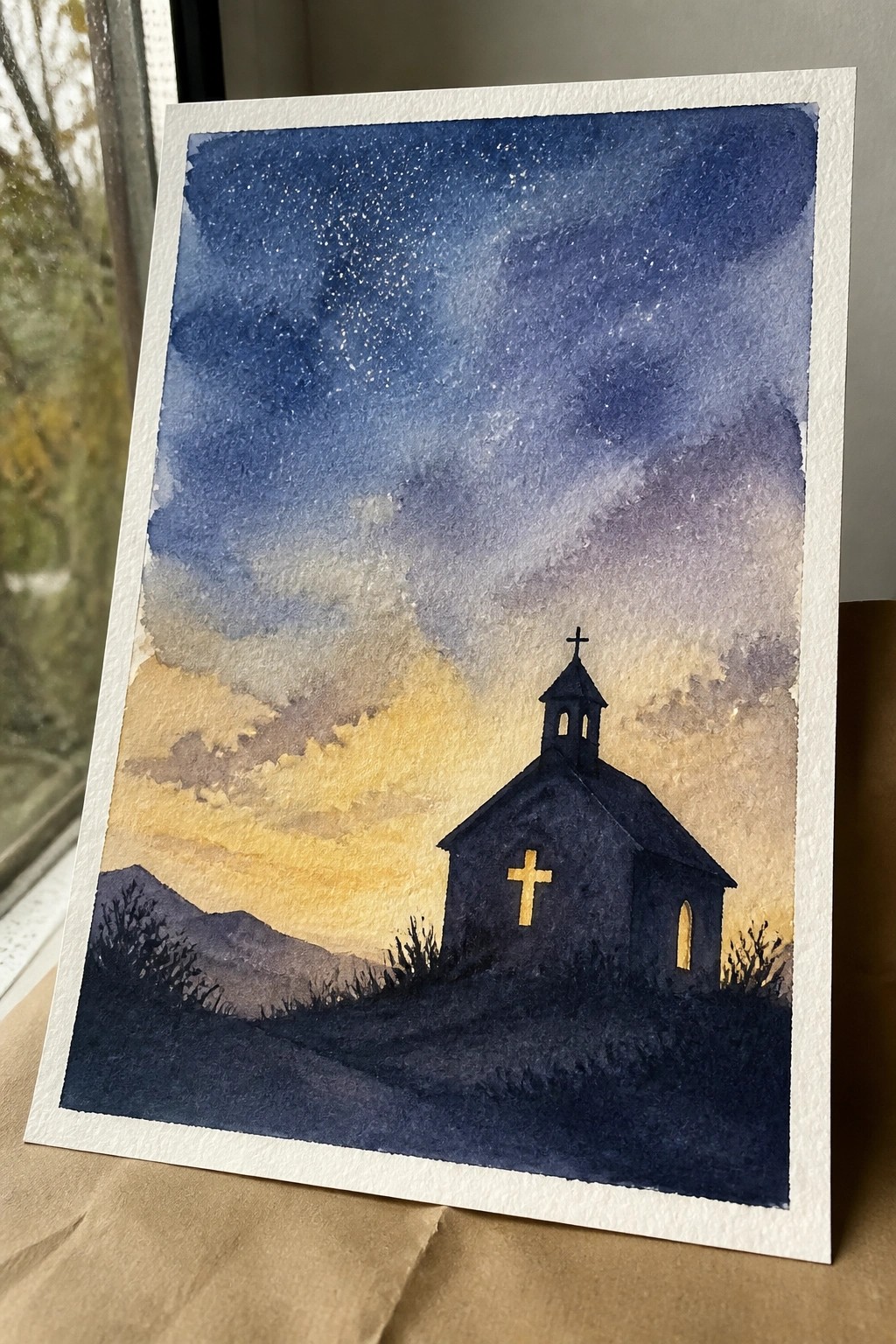 Watercolor silhouette of a church with glowing windows under a starry purple-orange twilight sky.
