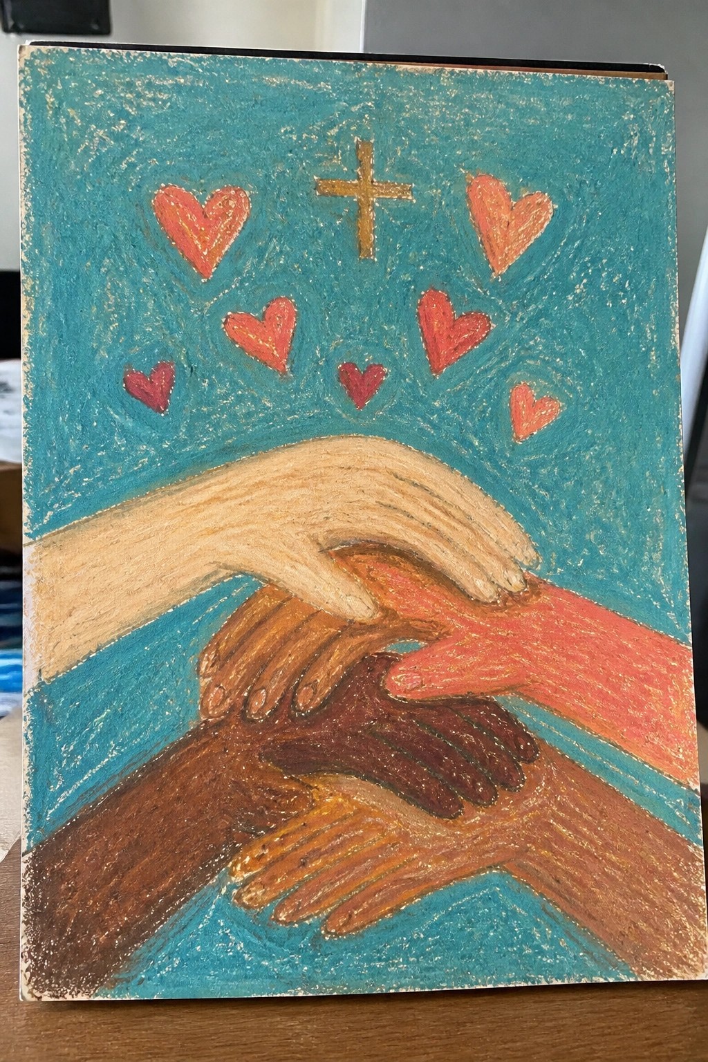 A painting of multiple hands with different skin tones clasped together, a gold cross above them, and red hearts on a teal background.