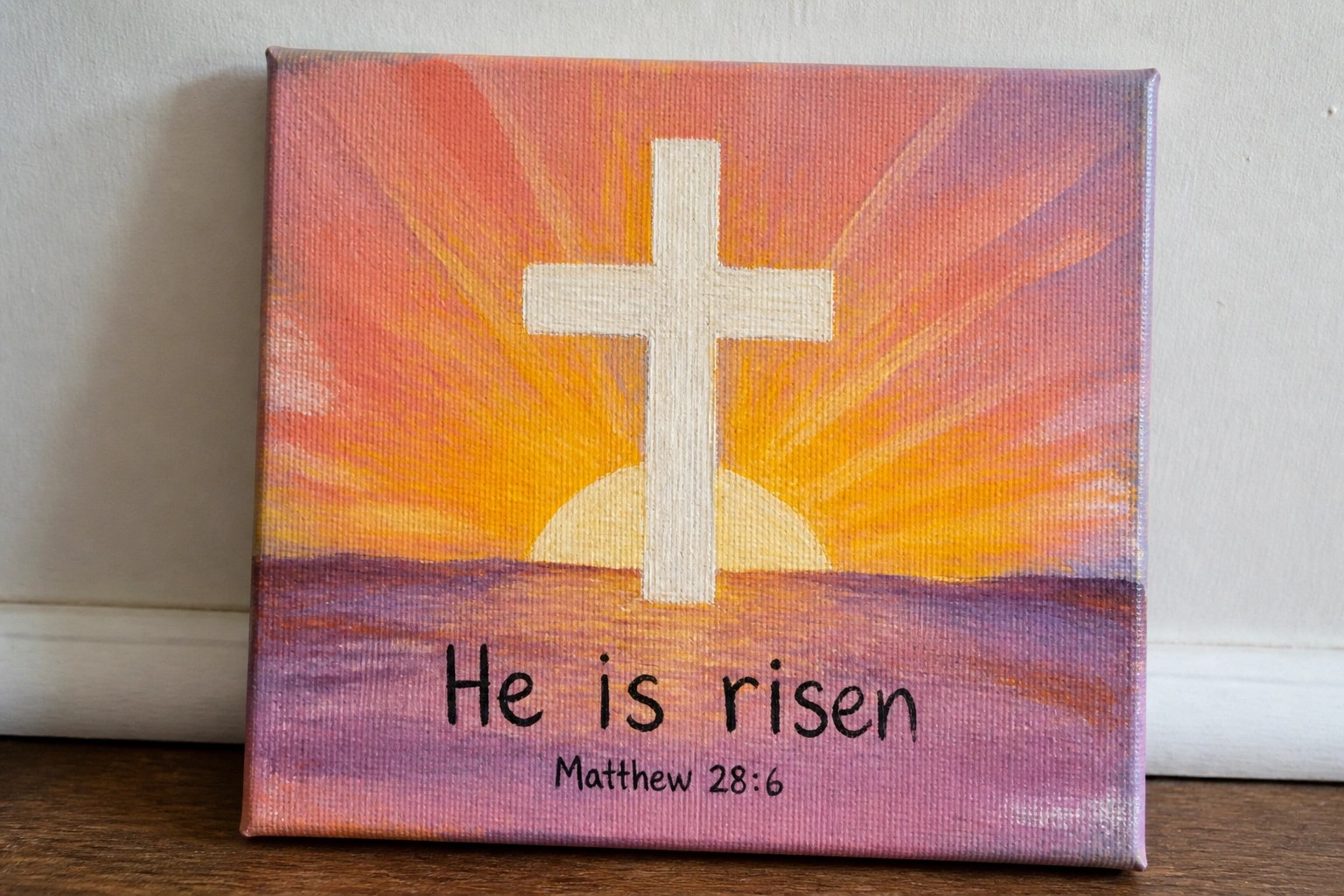 White cross on vibrant pink-orange sunset canvas with purple hills and "He is Risen" text
