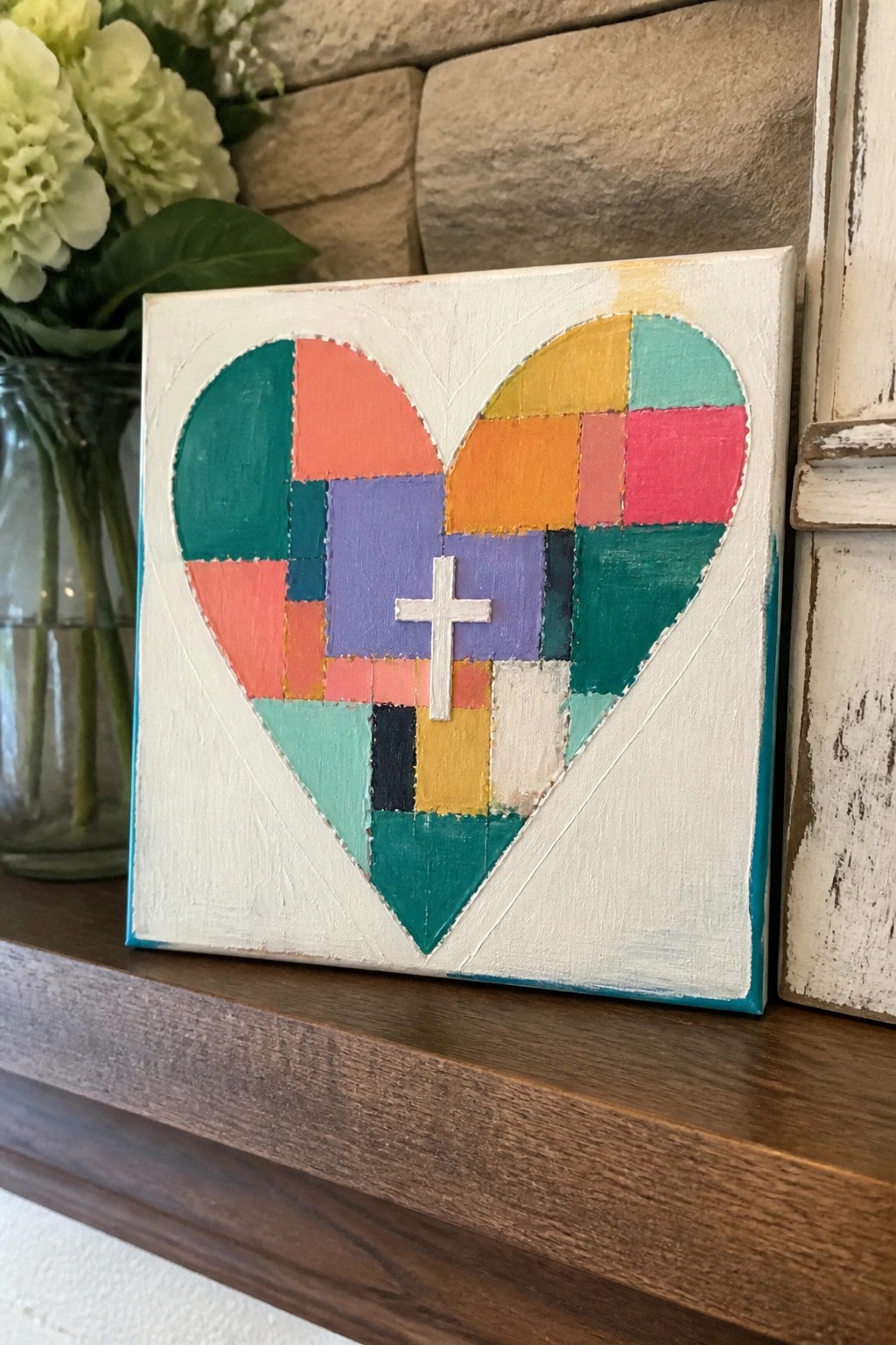 Colorful patchwork heart with white cross on canvas atop wooden mantel with flowers