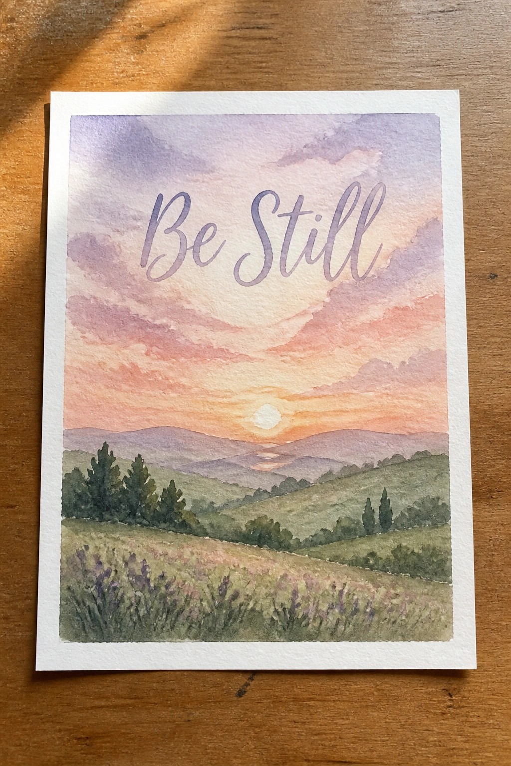 Watercolor sunset over rolling hills with purple flowers and Be Still text in sky
