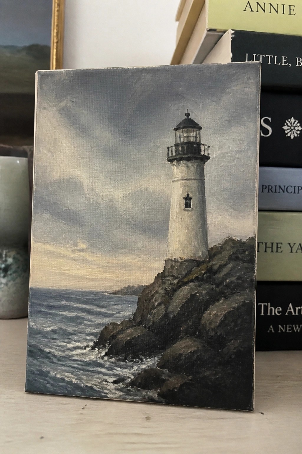 Miniature painting of white lighthouse on rocky cliff by crashing ocean waves beside books.