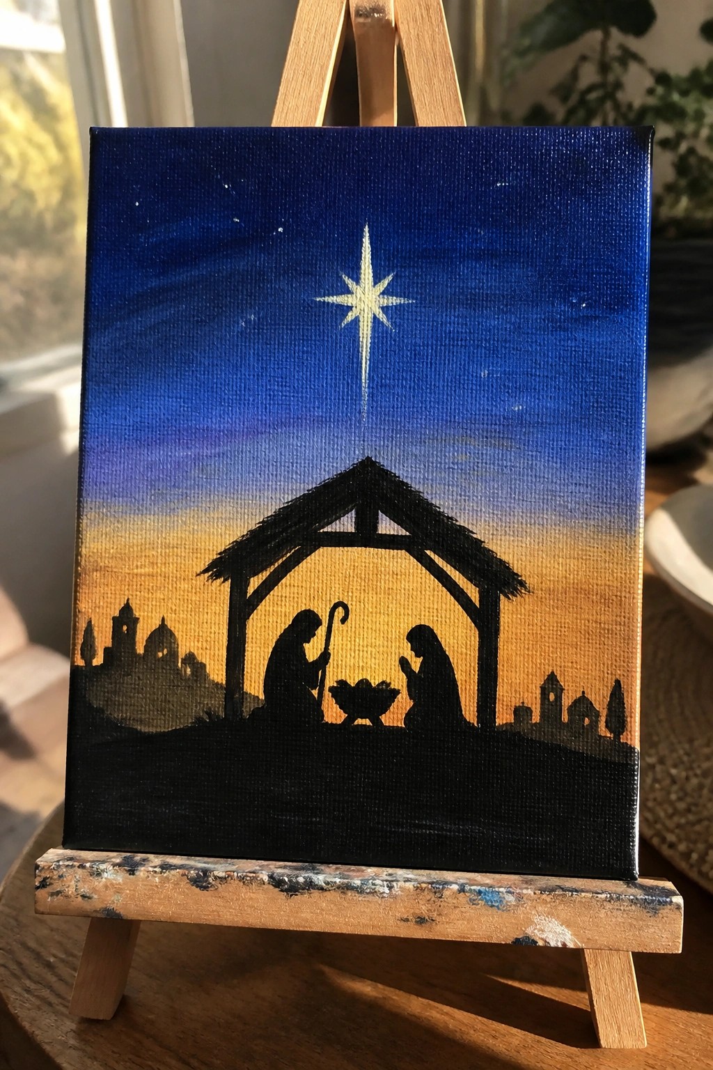 Silhouetted nativity scene with star above stable against blue-orange gradient sky on easel