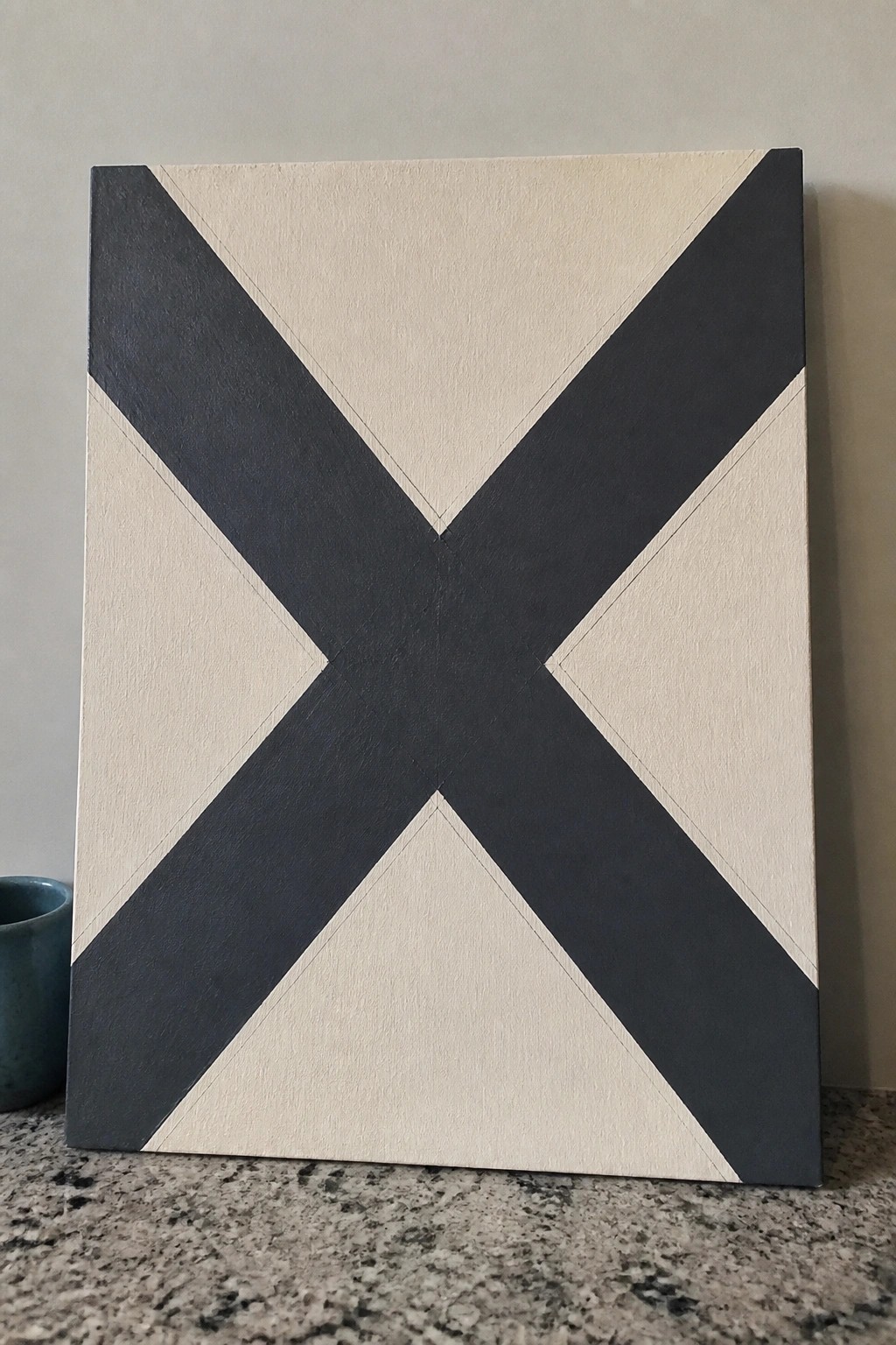 Square canvas with bold dark gray X on beige background atop granite counter