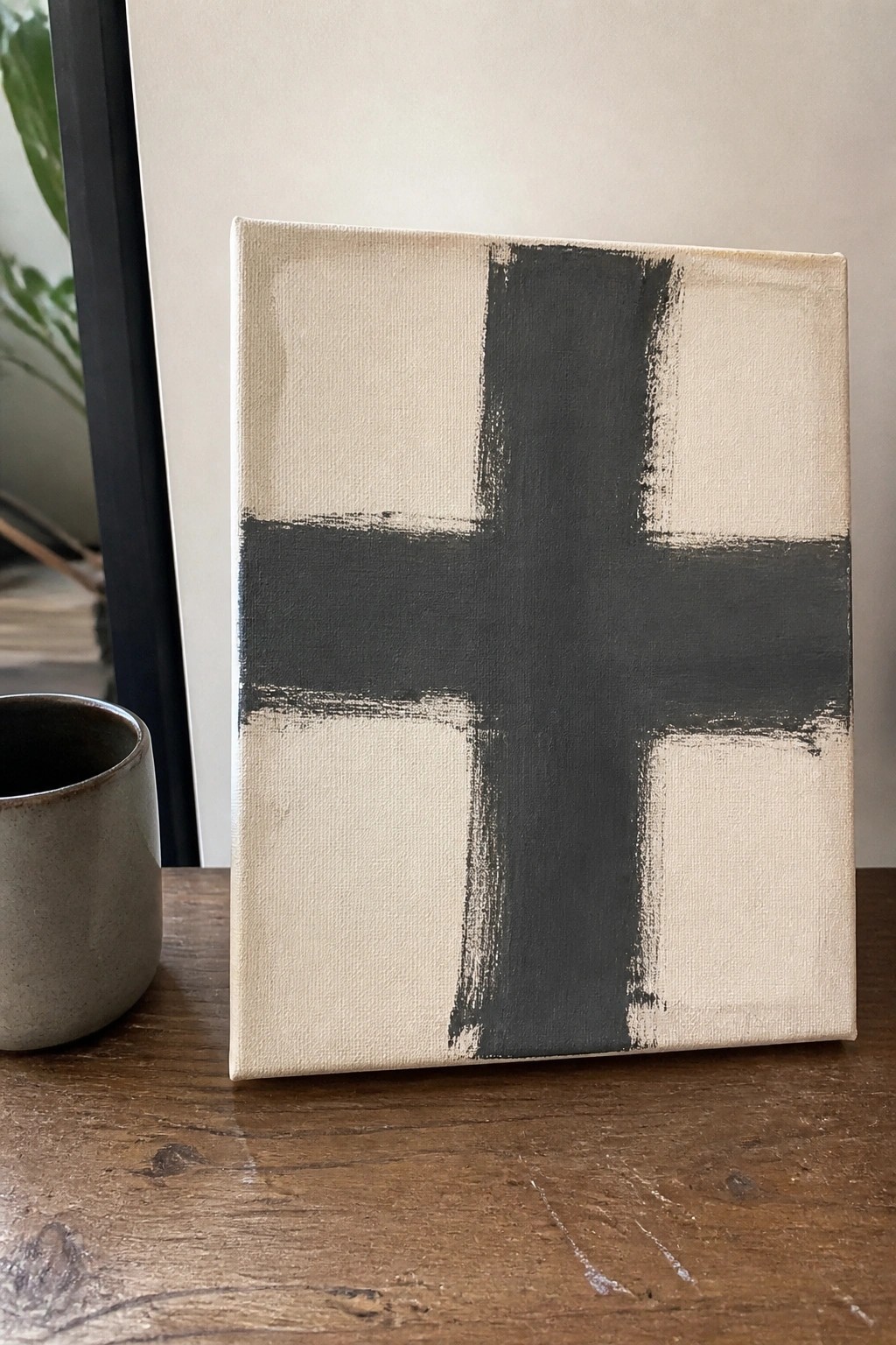 Beige canvas with bold gray cross brushstrokes beside a cup on wooden table.