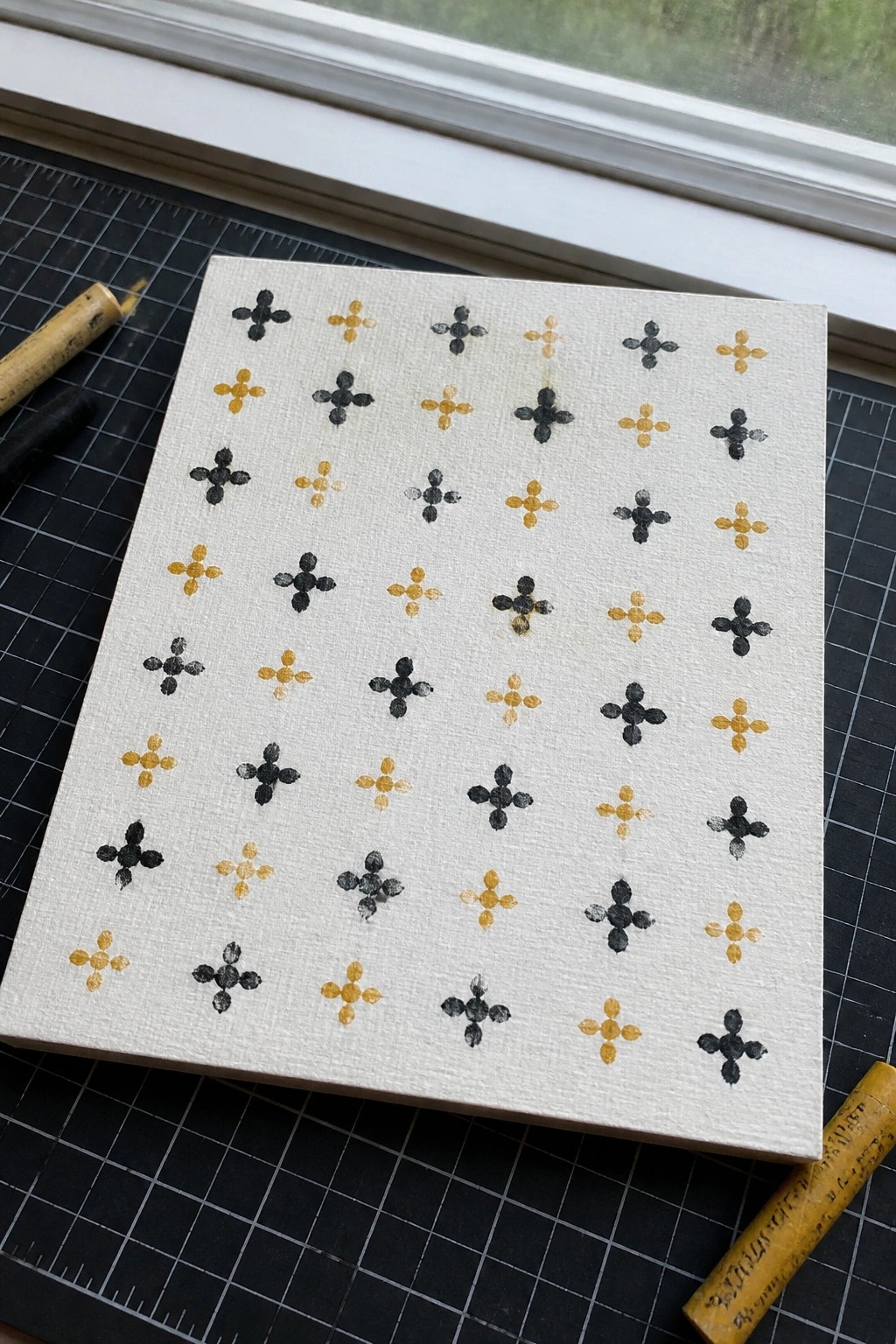 White canvas with rows of black and yellow cross patterns on gridded mat beside pastels.