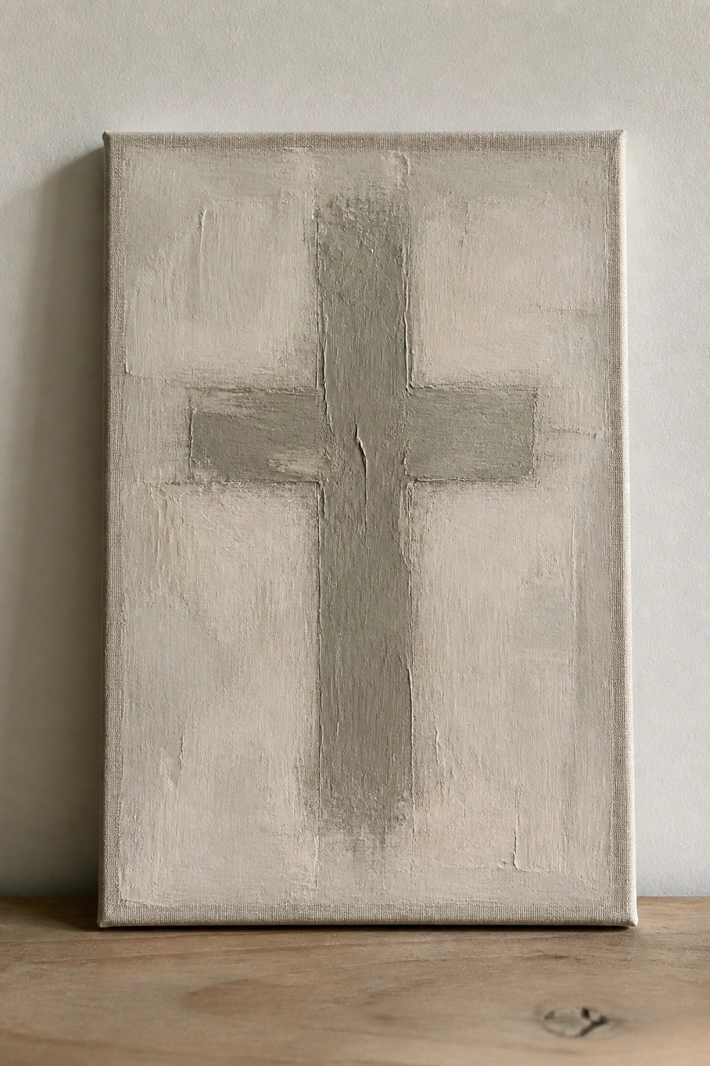 Textured minimalist painting of a gray cross on canvas against a white wall above wood.