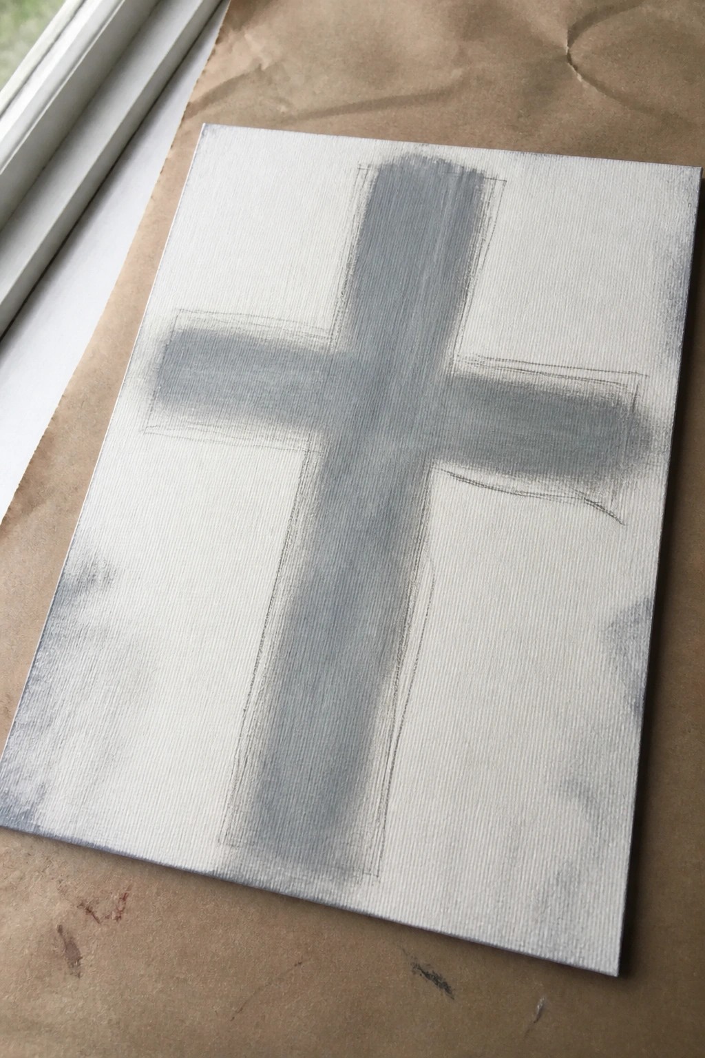 Gray cross spray-painted on white canvas with pencil outlines on brown paper