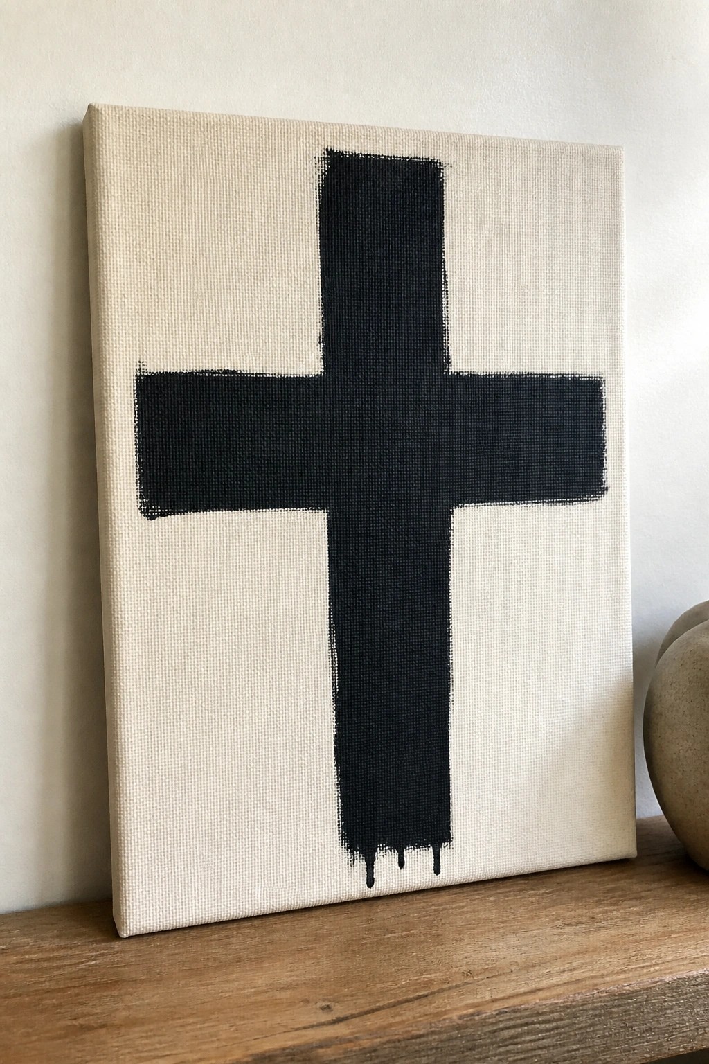 Black cross with drips on off-white canvas propped on wooden shelf beside vase