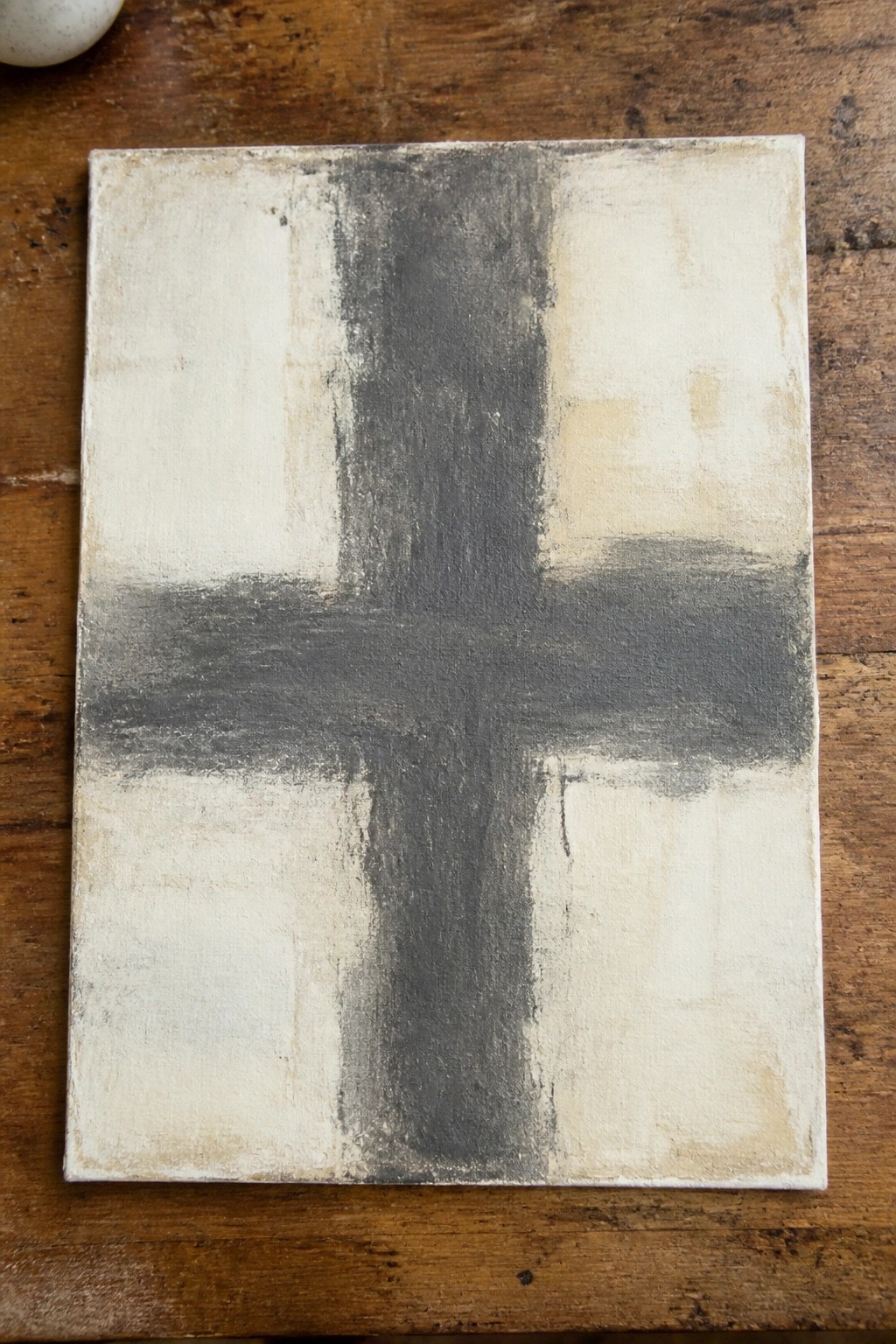 Abstract painting with a dark gray cross on textured off-white canvas atop wood.
