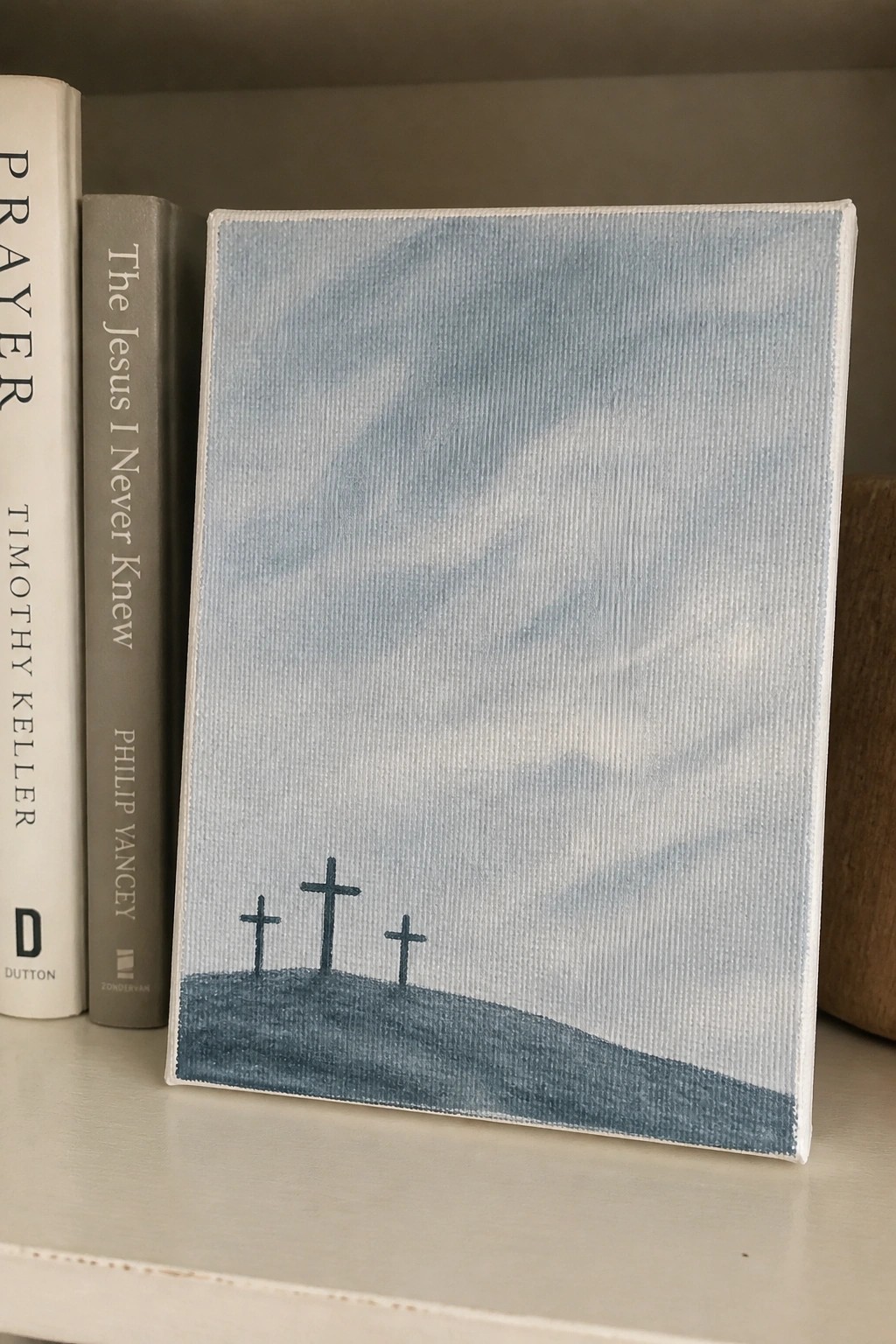 Canvas painting of three crosses on a hill next to two books on a shelf.
