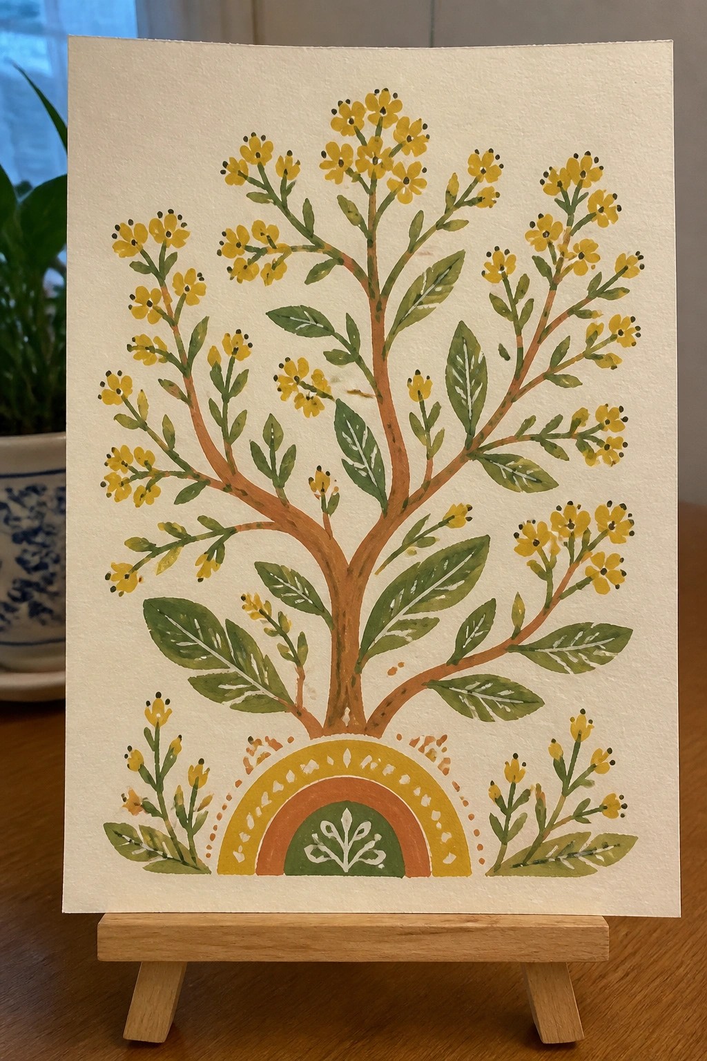 Watercolor painting of yellow-flowered tree with green leaves and rainbow arch on easel.