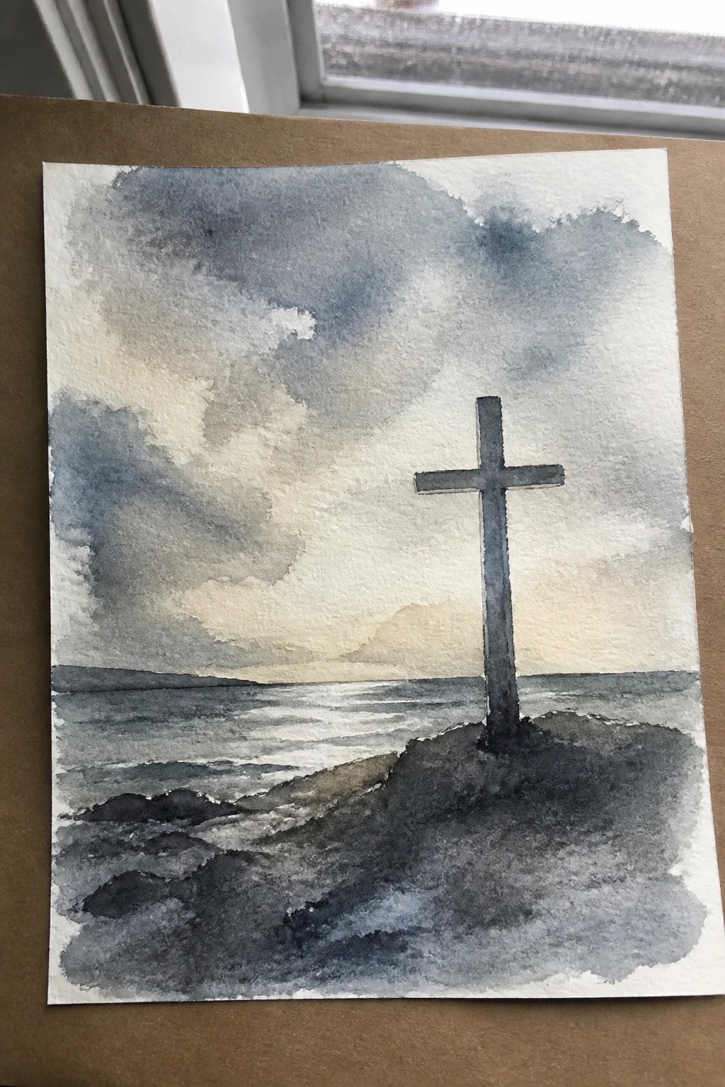 Watercolor of cross on rocky shore by sea under dramatic cloudy skies at sunset