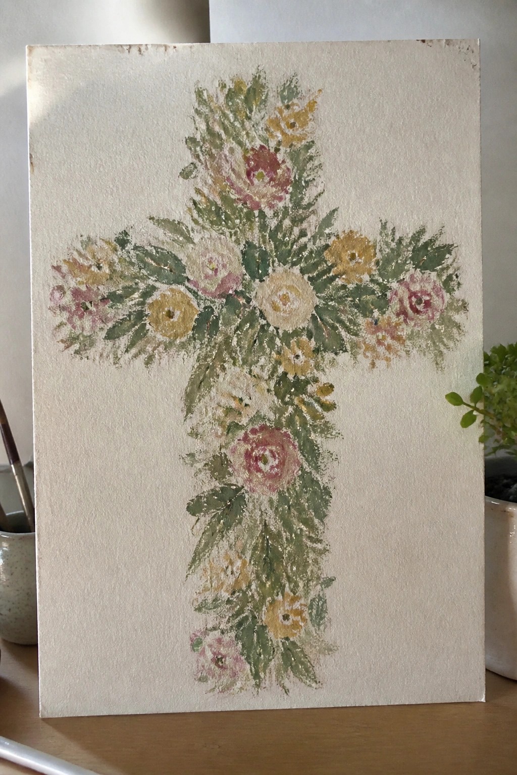 A painting of a cross covered in clusters of pink, yellow, and white flowers with green leaves on a textured off-white surface.