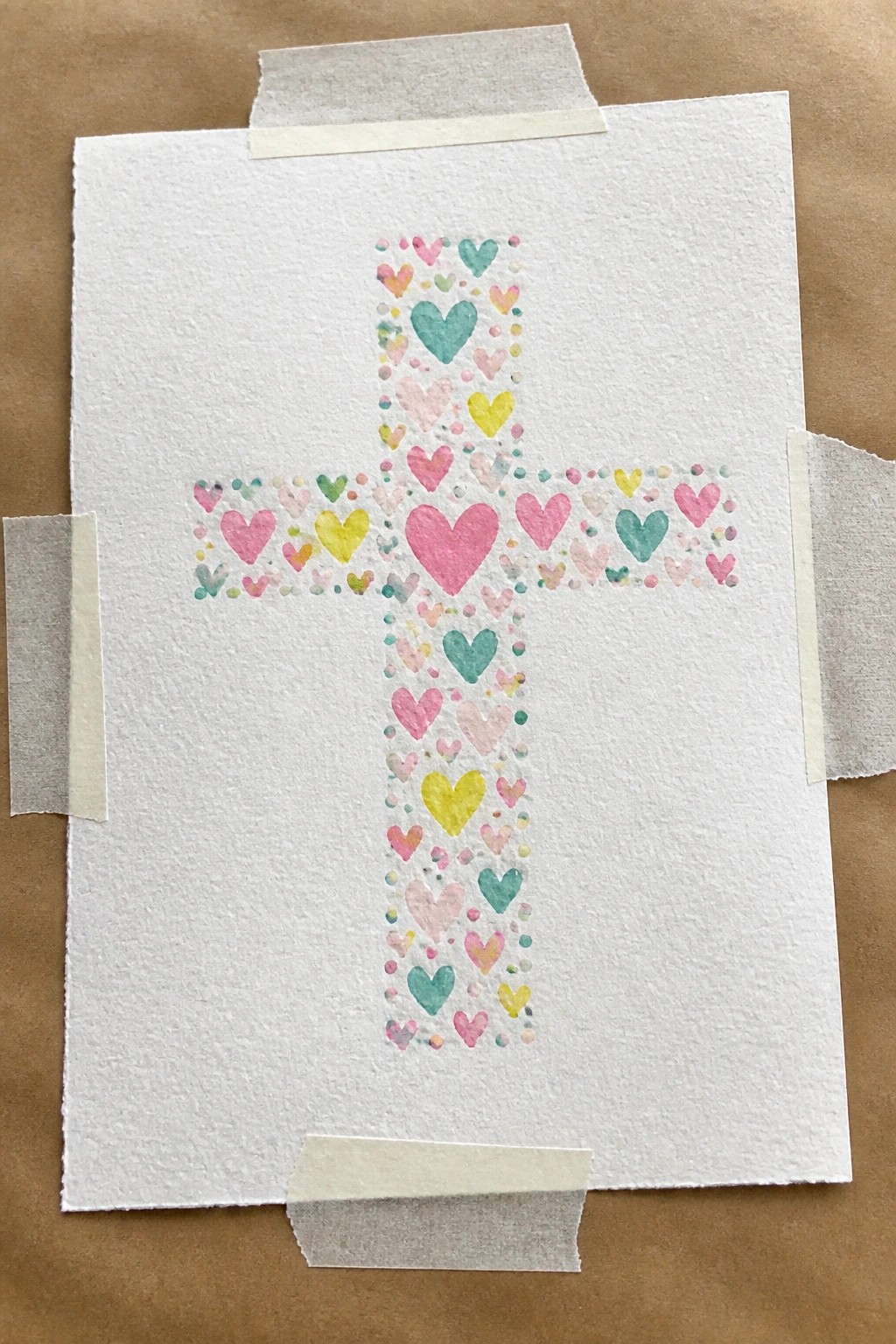 Watercolor cross of colorful hearts on white paper taped to brown surface