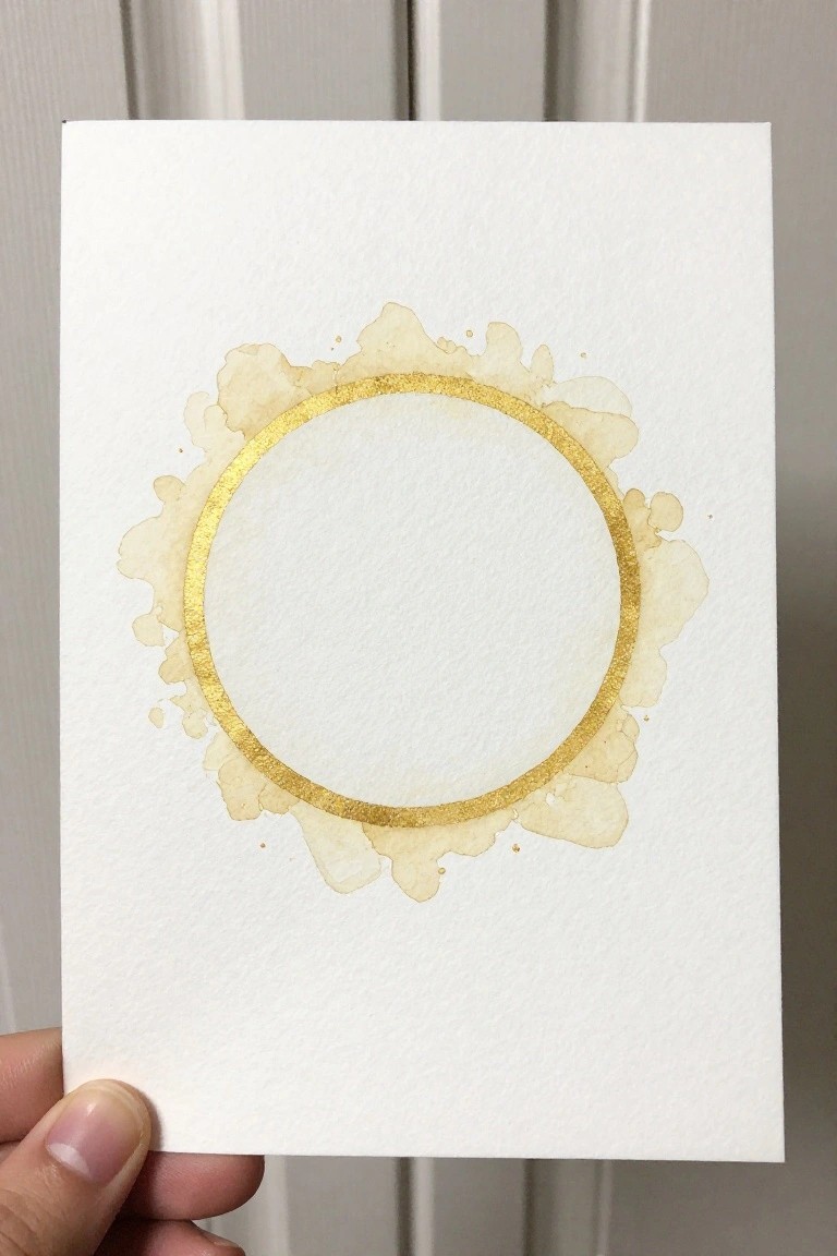 Hand holding white card with gold circle and beige watercolor splashes around it