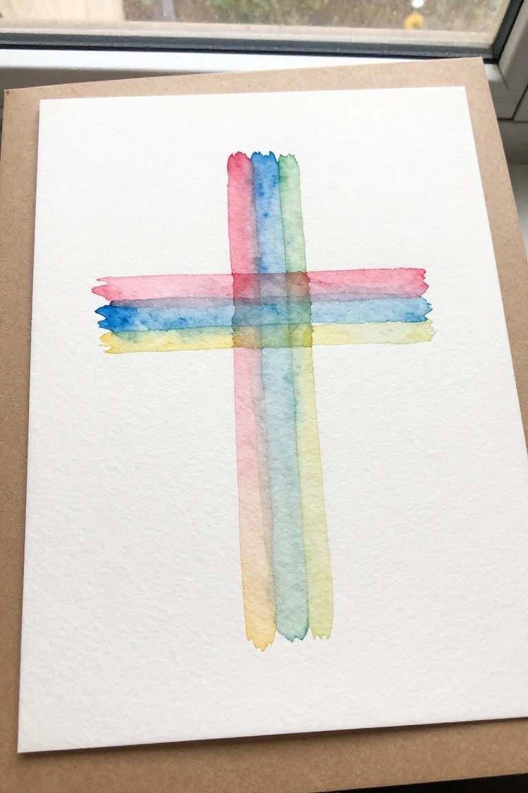 Watercolor cross in pink, blue, green, and yellow on white paper.