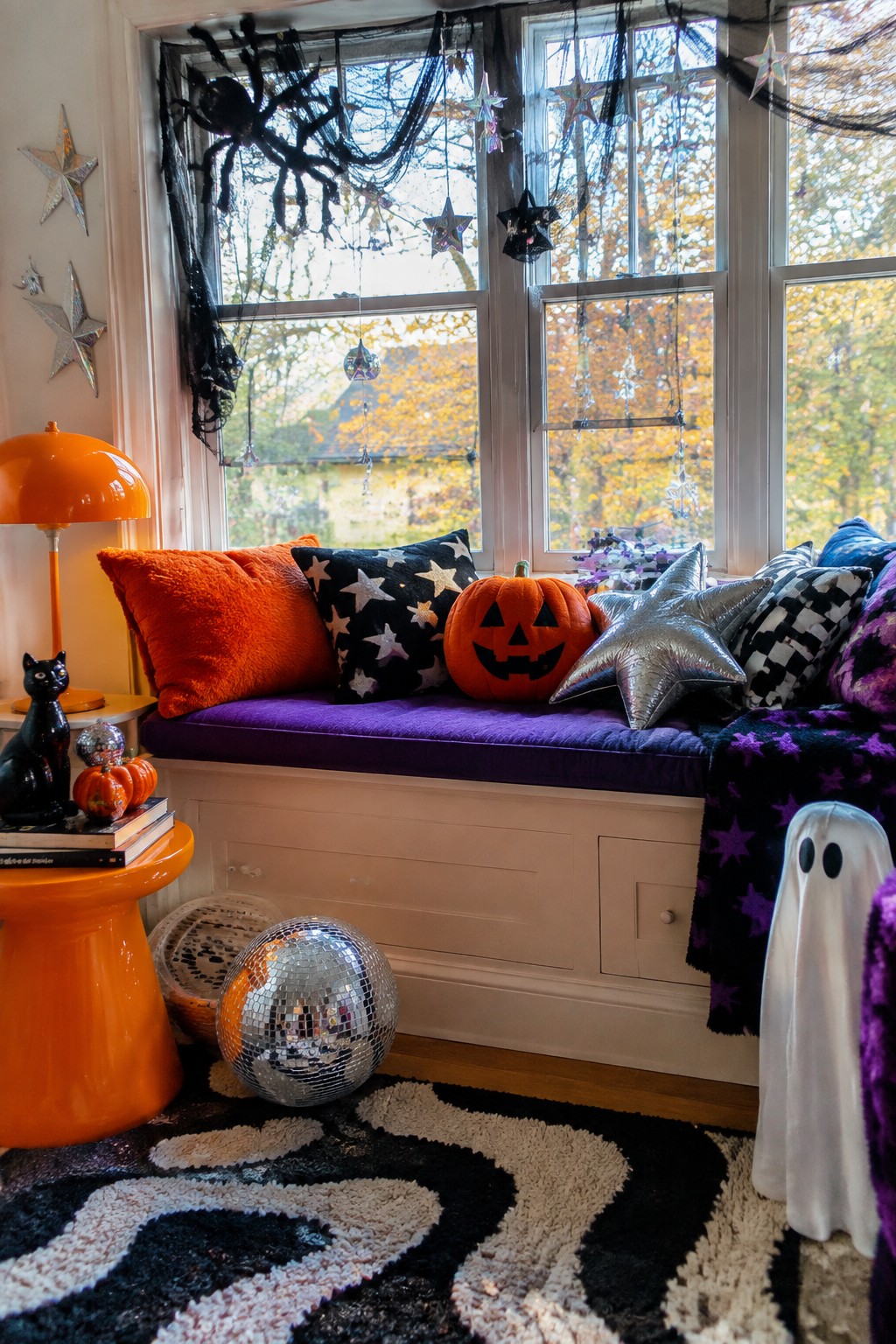 Window seat with purple cushion, carved jack-o'-lantern, star pillows, large black spider in a web, hanging stars, disco ball, and a white ghost figure on the floor.