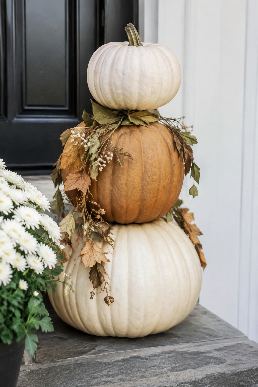 Stacked white, orange, and white pumpkins with autumn leaves beside white flowers