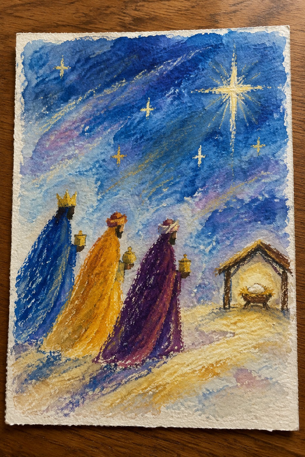 Three robed figures in blue, yellow and purple carry lanterns toward a manger under bright star in watercolor.