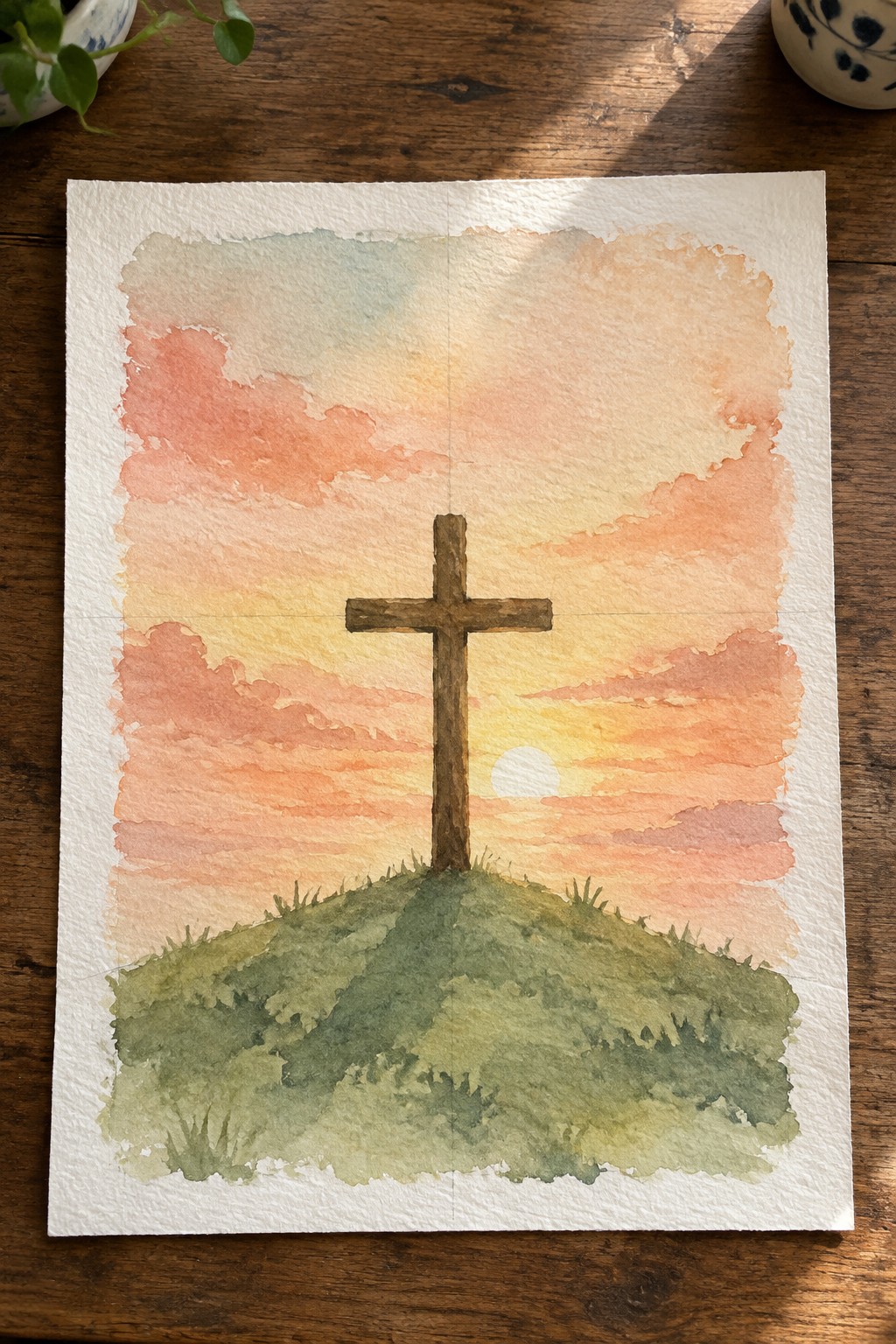 Watercolor of wooden cross on grassy hill under pink orange sunset sky with clouds.