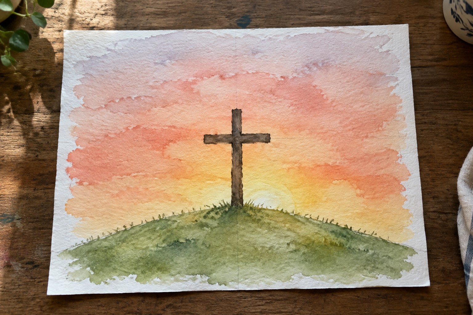 Watercolor of wooden cross on grassy hill under pink orange sunset sky with clouds.