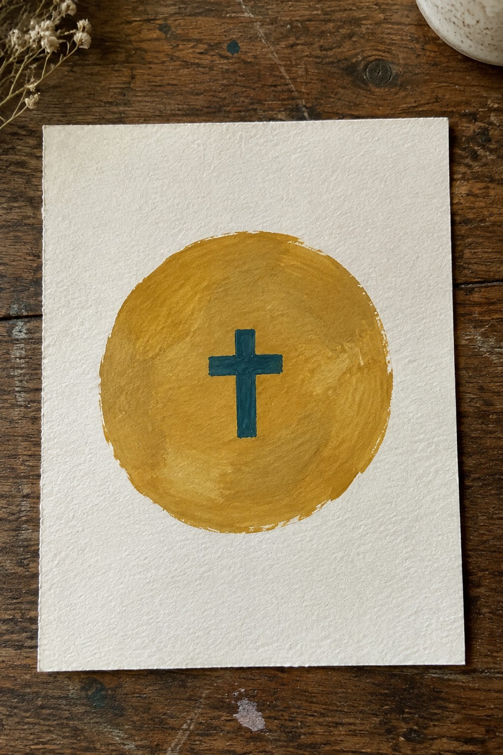 Yellow circle with teal cross painted on white paper over wood