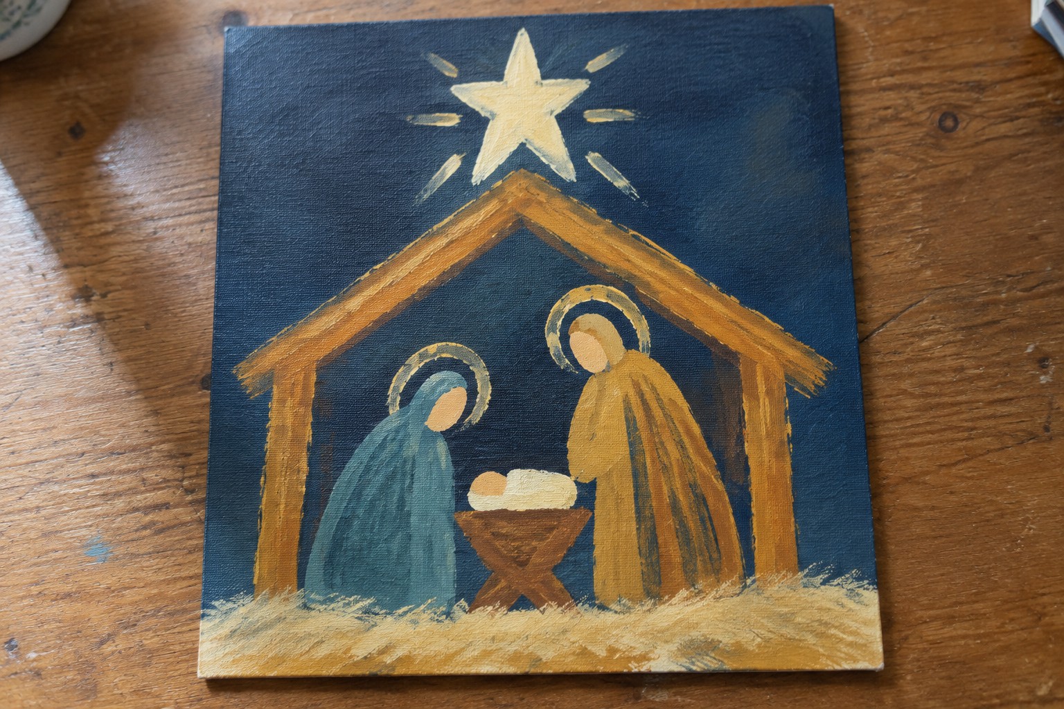 Nativity painting with blue-robed Mary, brown-robed Joseph, and haloed infant under golden star.