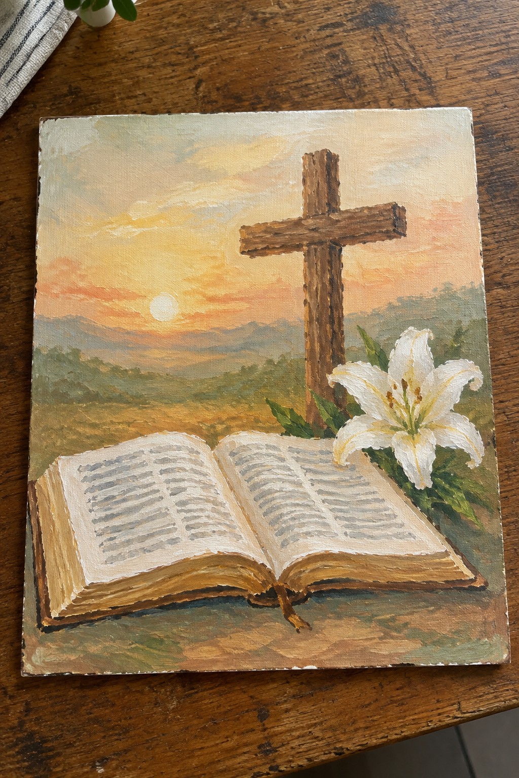 Open Bible, wooden cross and white lily against orange sunset over green hills.