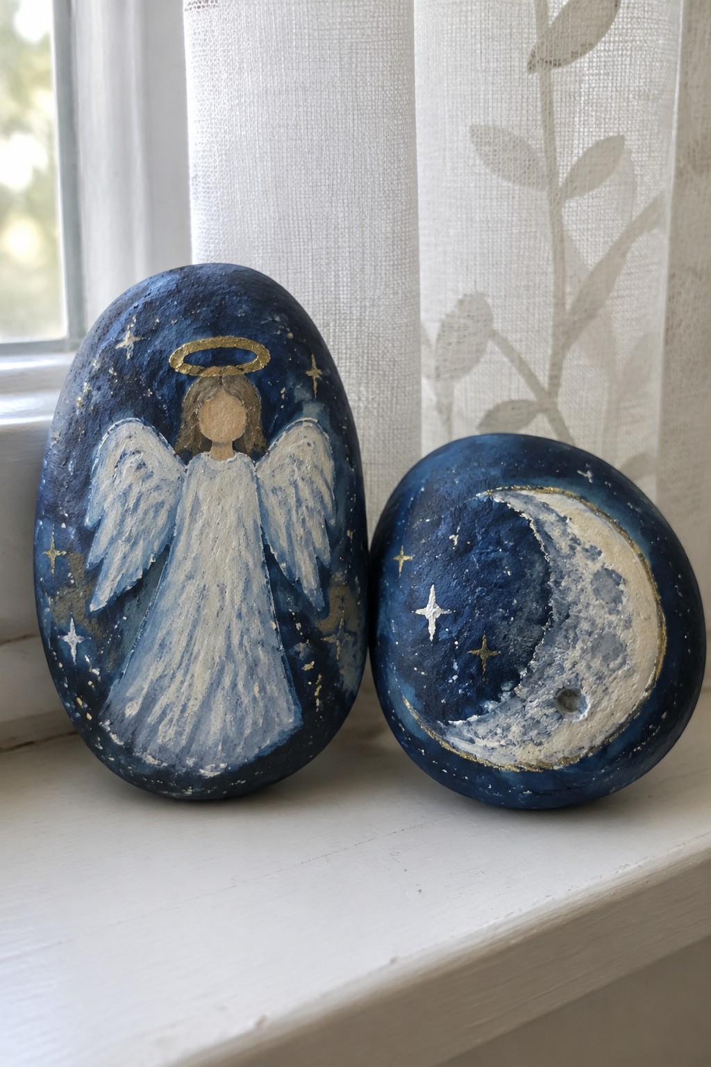 Two blue rocks painted with an angel and starry crescent moon in gold and white.